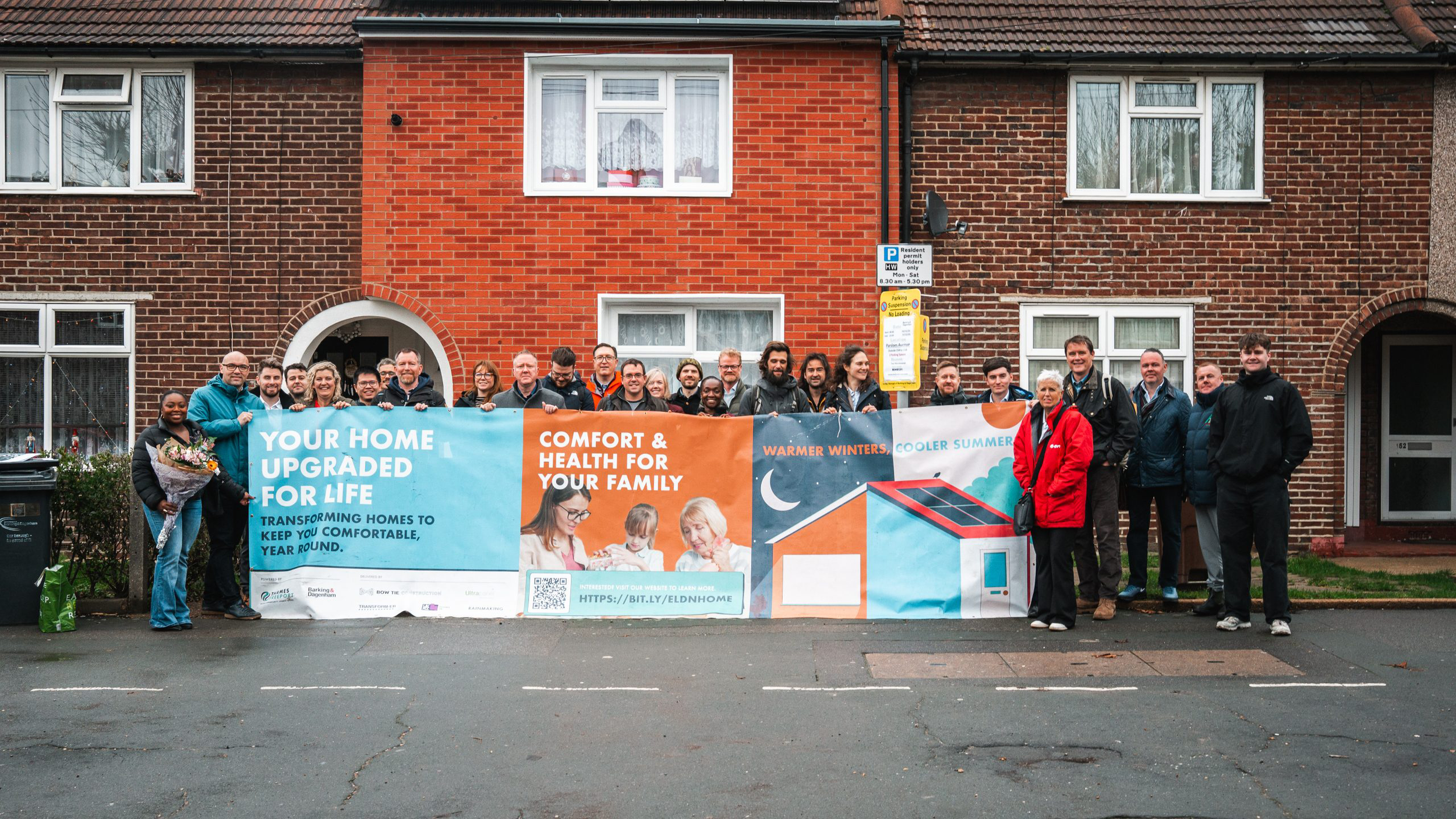 Group of people standing outdoors holding a large banner promoting home comfort and energy efficiency in front of a brick residential building.