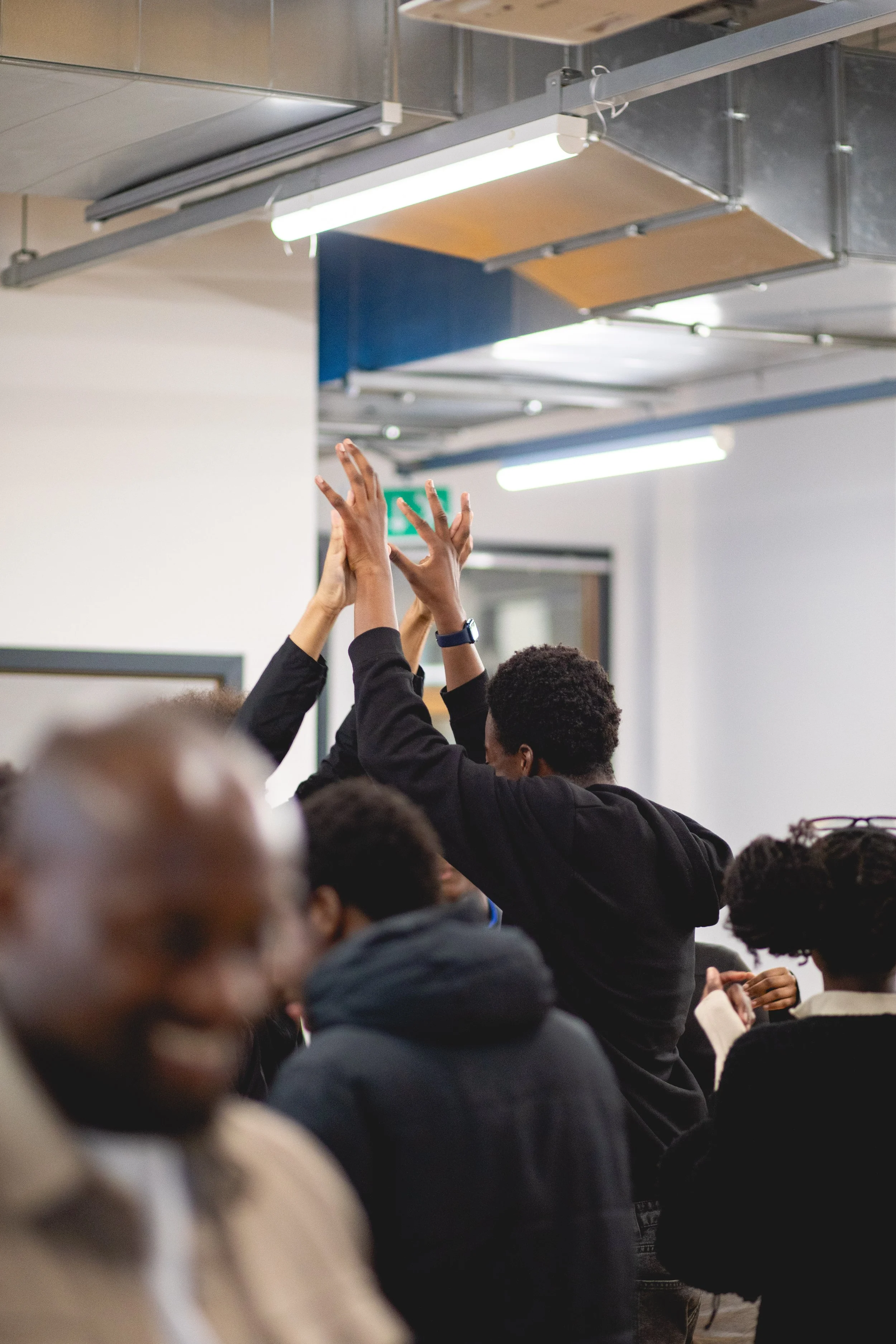 People raising their hands during a meeting or seminar in a classroom or conference room with fluorescent lighting and white walls.