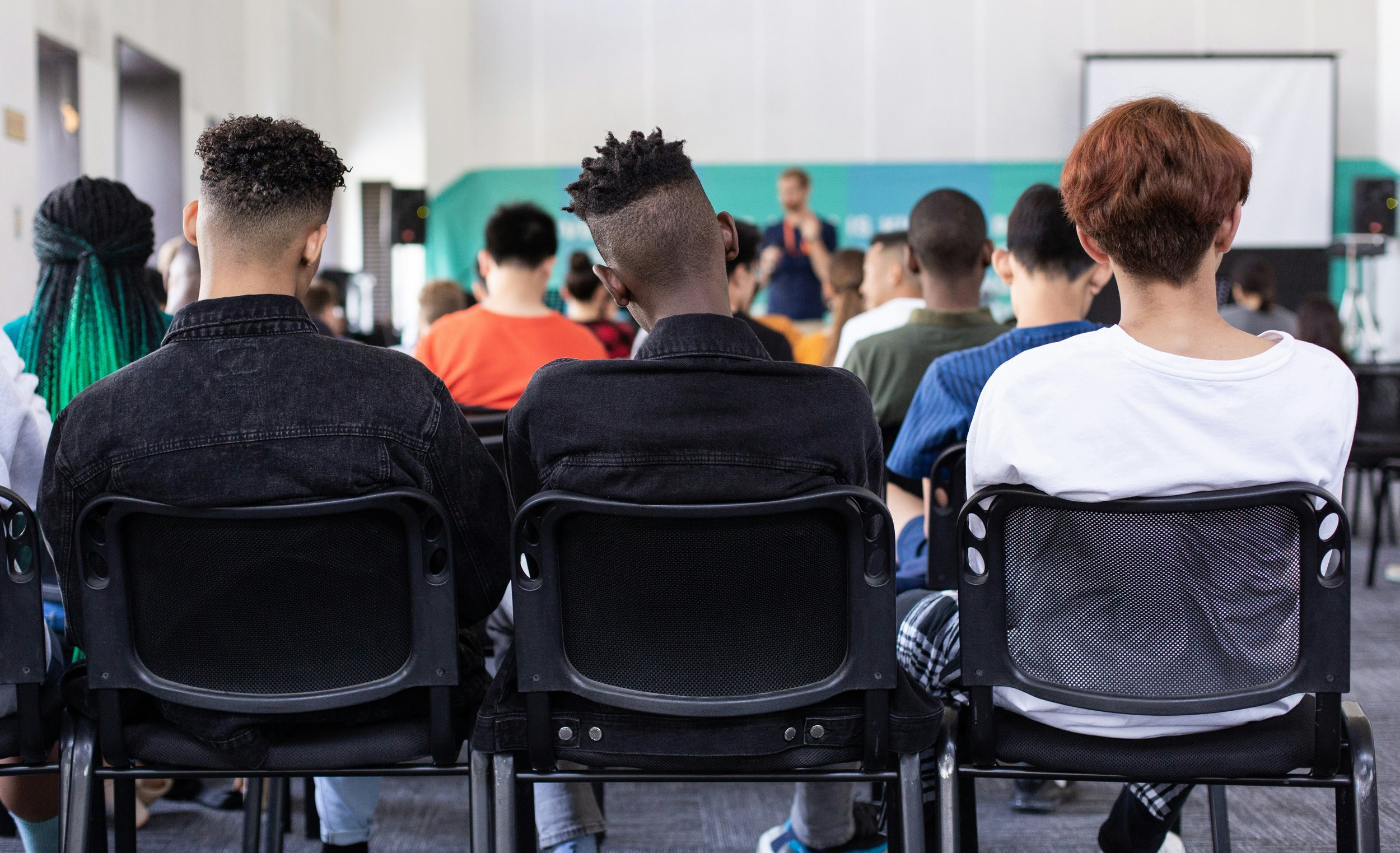 A group of students sitting on black chairs in a classroom, facing a teacher at the front of the room, with some students taking notes and others listening.