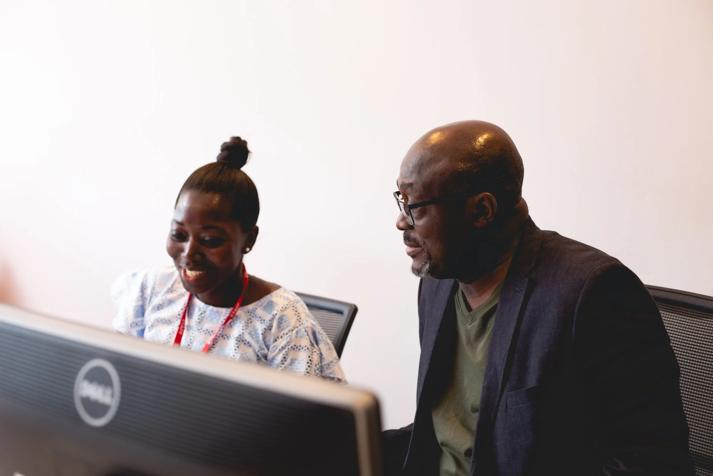 A young woman and an older man sit together at a desk, looking at a computer screen and engaging in a discussion in an office setting.