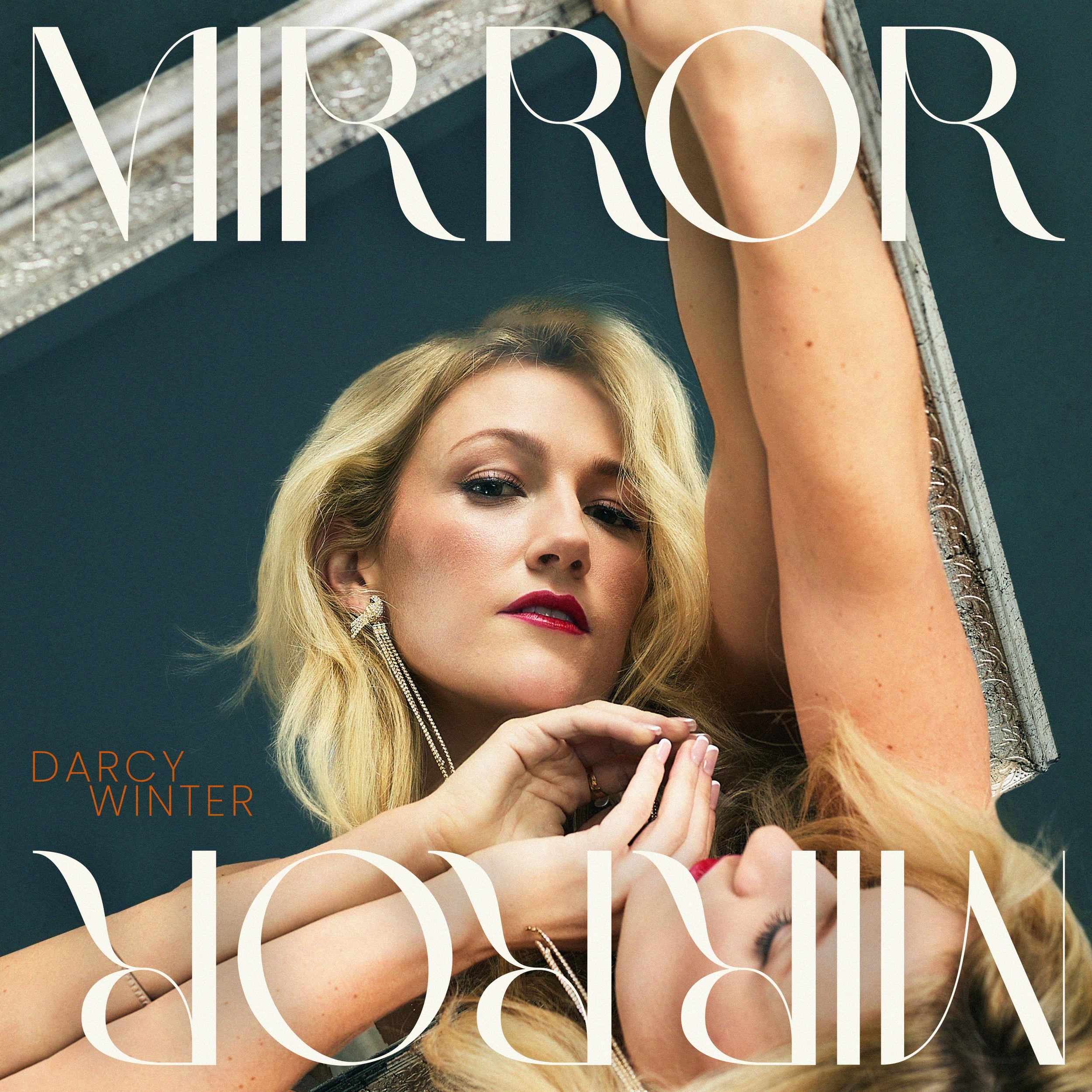 A close-up of Darcy Winter, wearing earrings and makeup, posed with her arm over her head. The background is dark green, and there is white text overlay that reads "MIRROR MIRROR" and "DARCY WINTER".