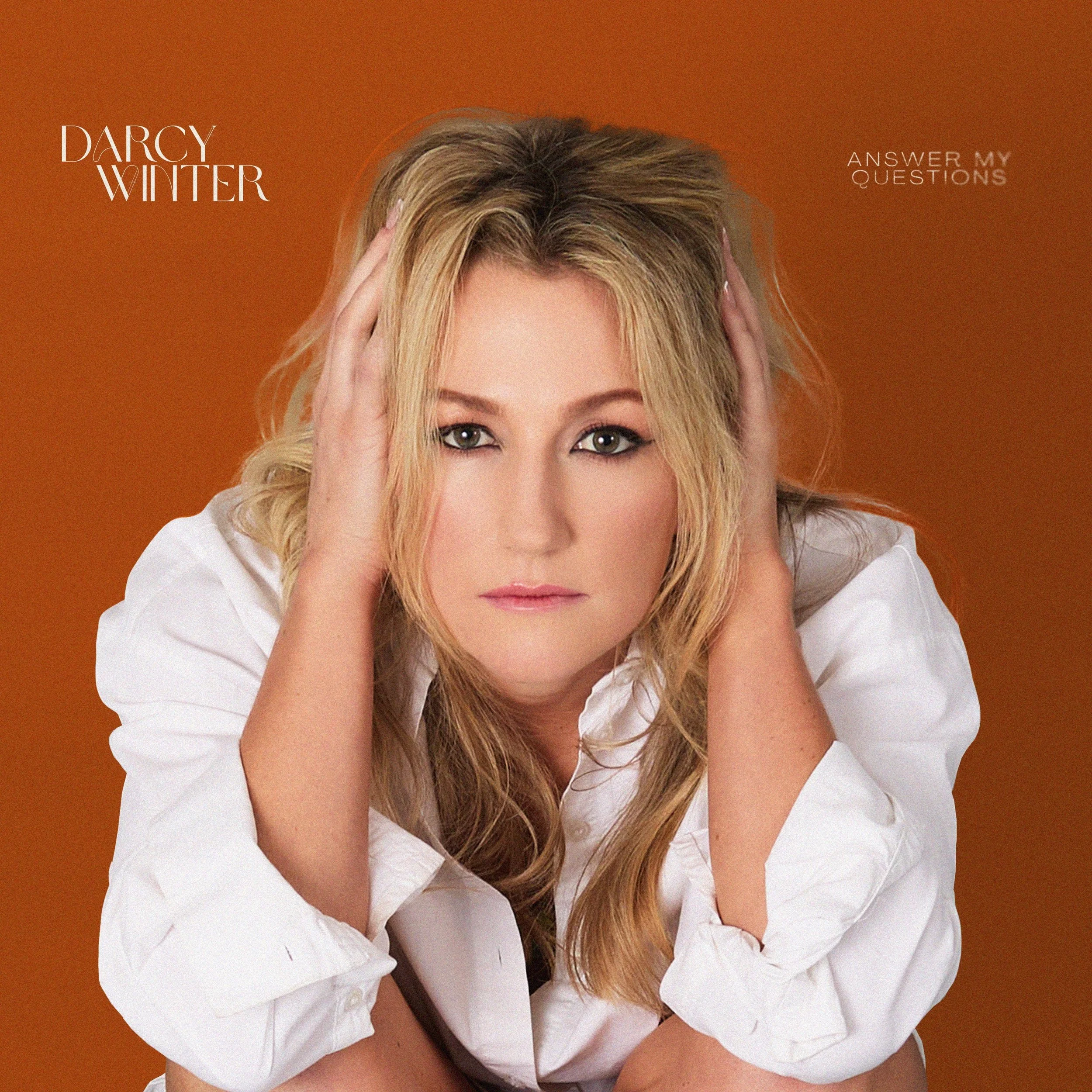 Close-up of a Darcy Winter in a white shirt, resting her head on her hands against an orange background, with the text 'DARCY WINTER' on the left and 'ANSWER MY QUESTIONS' on the right.