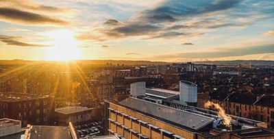 Sunrise over a city skyline with buildings, some emitting smoke, under a partly cloudy sky.