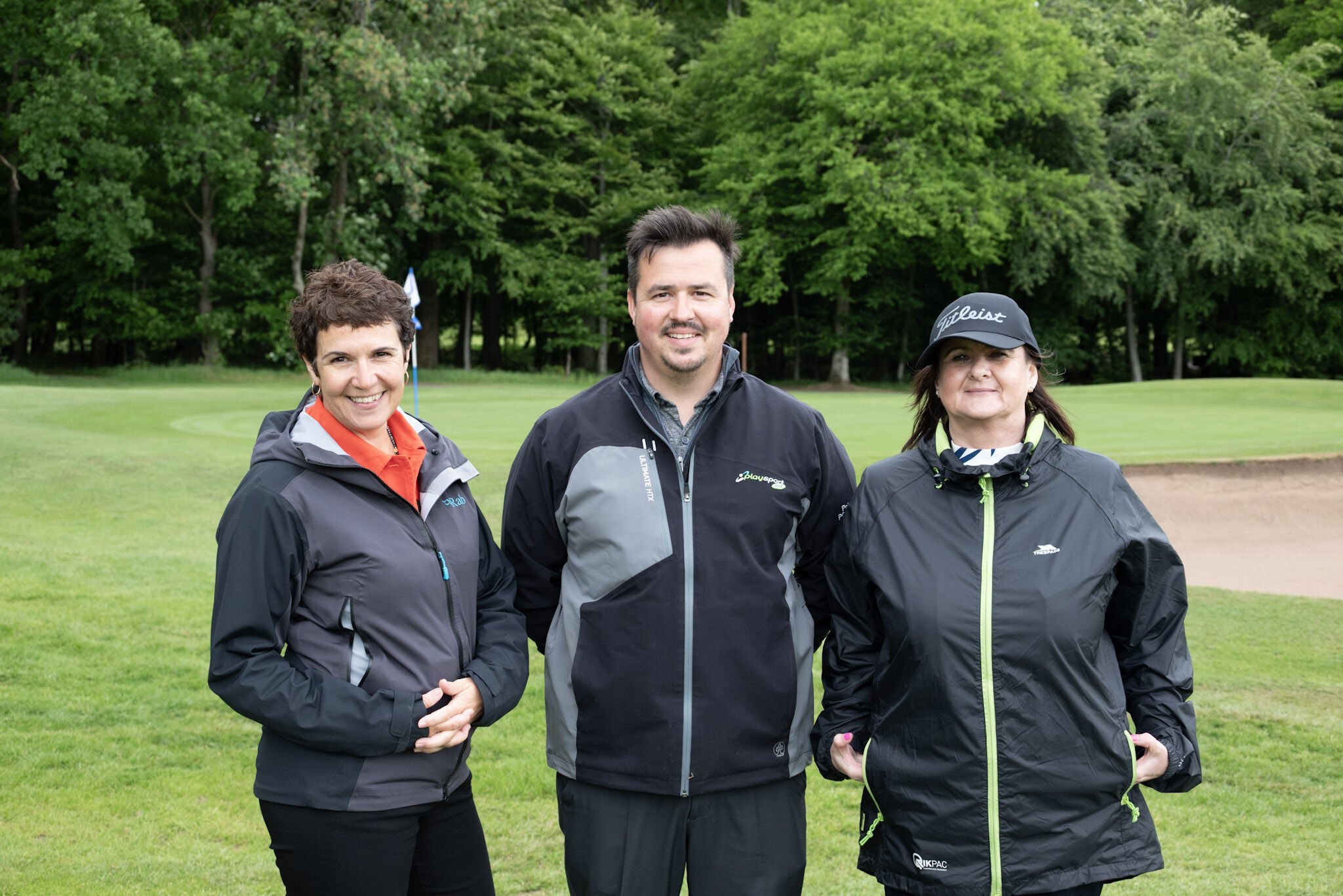 Three people standing on a golf course with trees in the background, wearing jackets and sporting golf apparel.