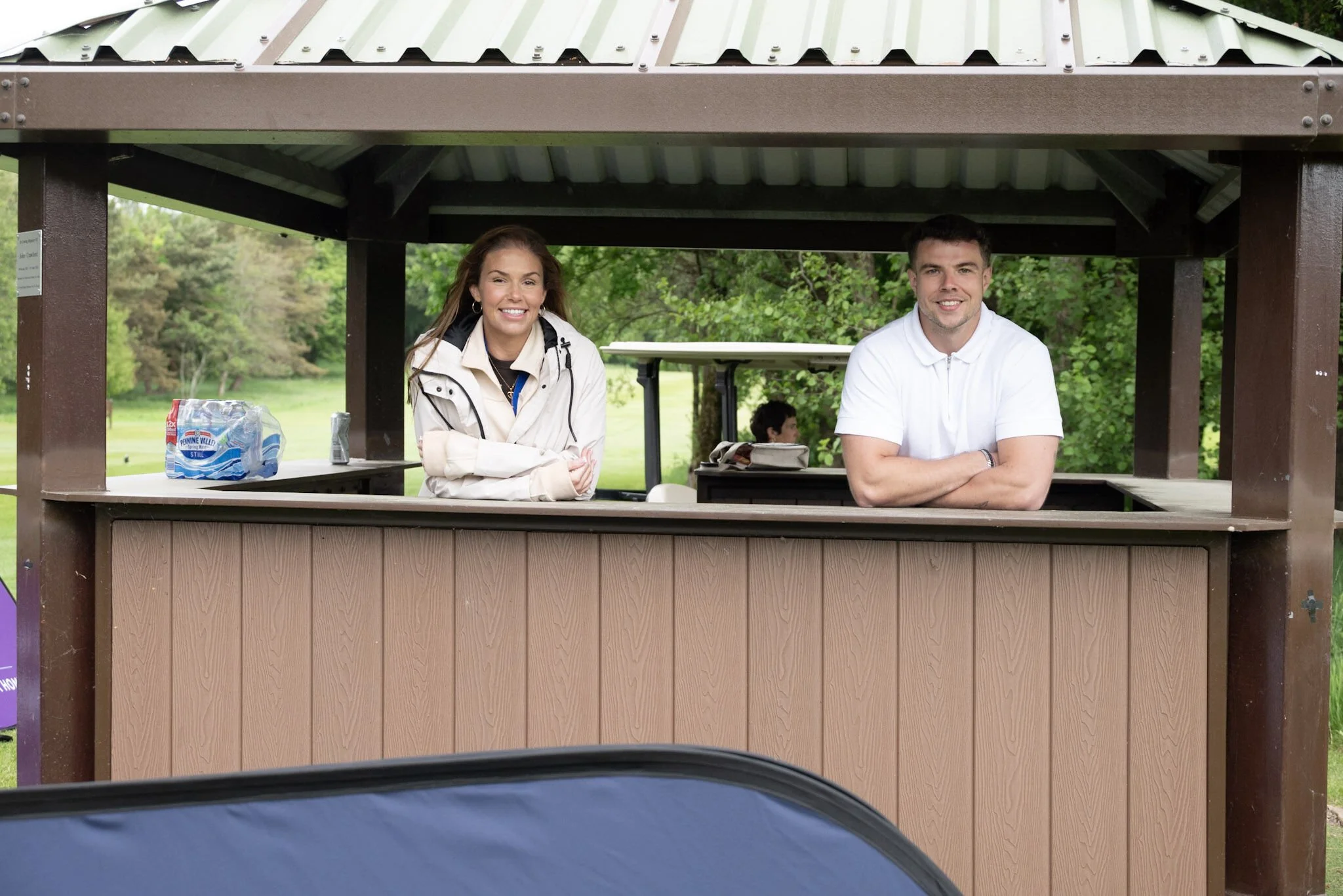 Two people, a woman and a man, are standing behind a wooden counter at an outdoor pavilion on a golf course, with trees and grass in the background.