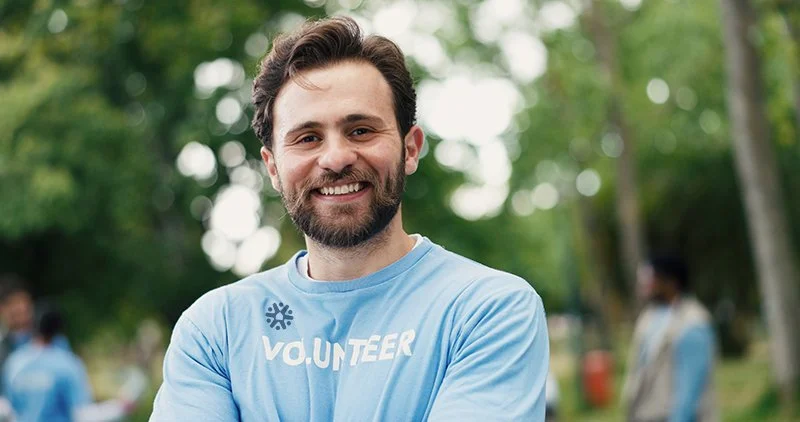 Smiling man with beard wearing a blue volunteer shirt outdoors with trees and other people in the background.