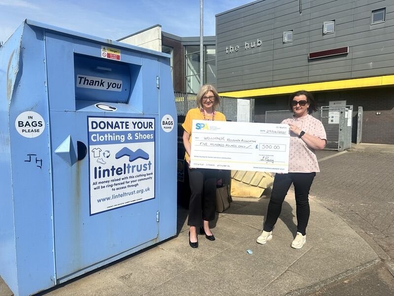 Two women standing outside next to a blue clothing donation bin, holding a large check for 500 pounds donated to a homeless advocacy organization.