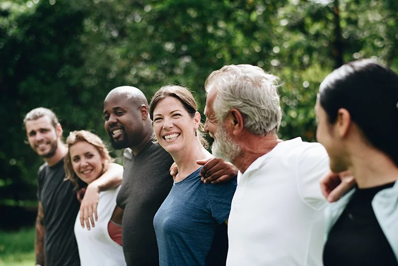Group of diverse people standing outdoors, smiling, with arm around each other's shoulders, in a green park.
