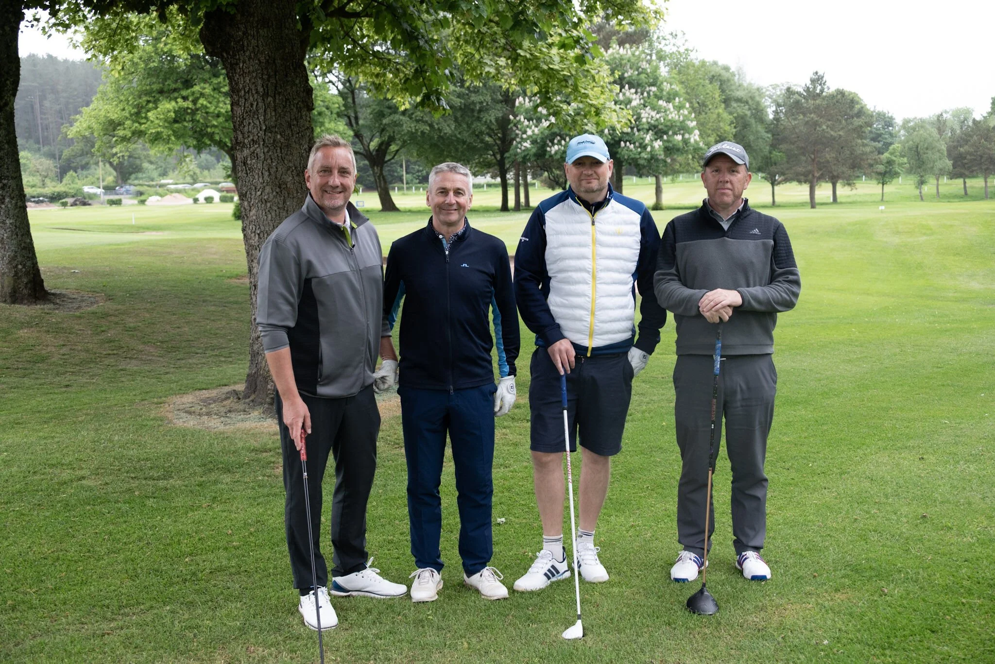 Four men standing together on a golf course, holding golf clubs with trees and a grassy landscape in the background.