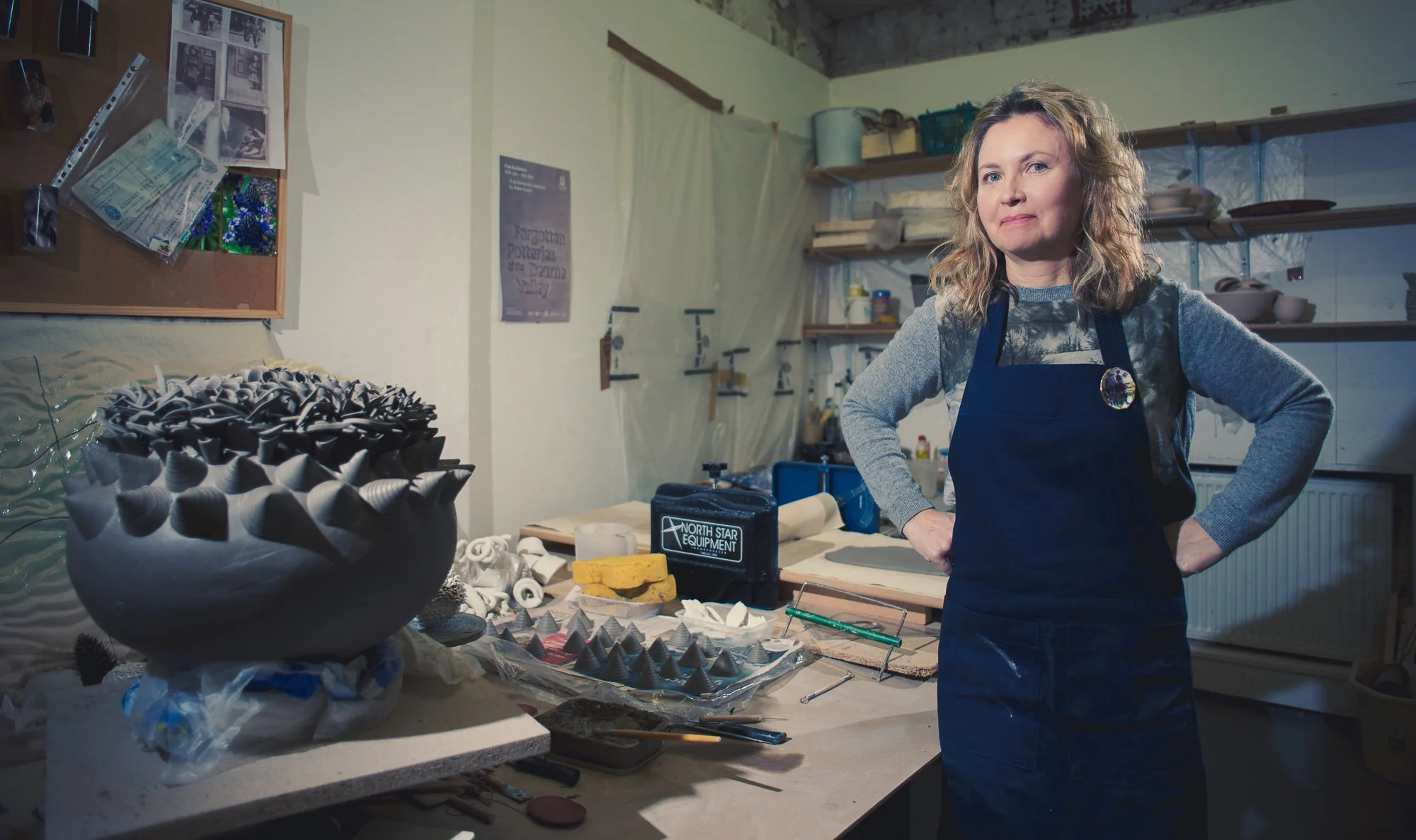 A woman with curly blonde hair wearing a gray sweater and black apron stands in a pottery studio with her hands on her hips, surrounded by pottery tools and ceramic pieces.