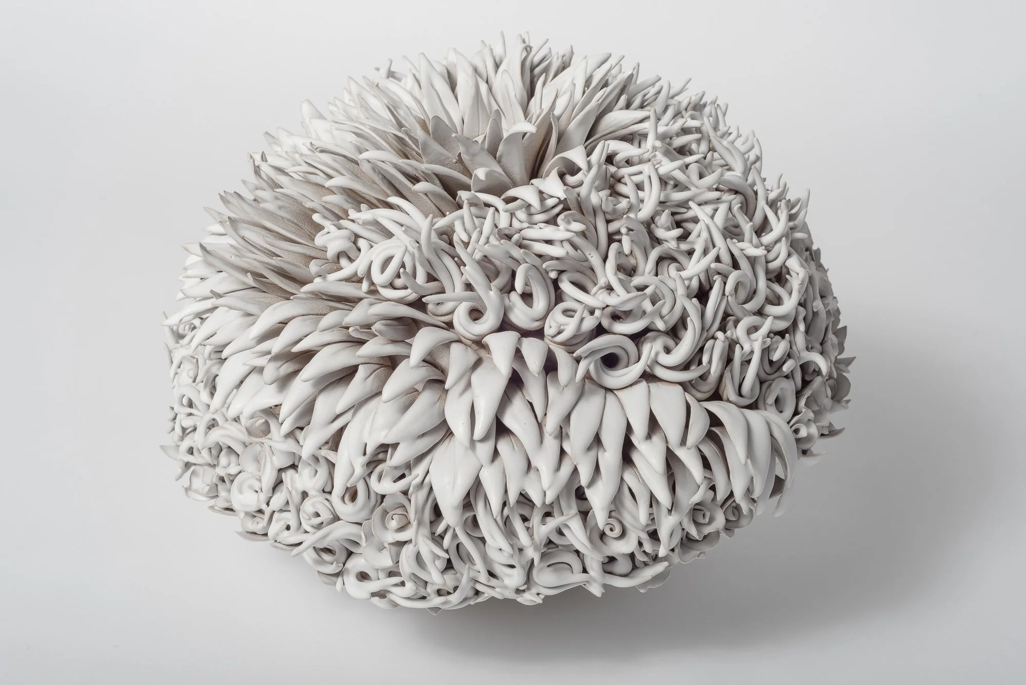 Close-up of a spherical object made of intricately curled and twisted white ceramic or clay pieces on a plain light gray background.