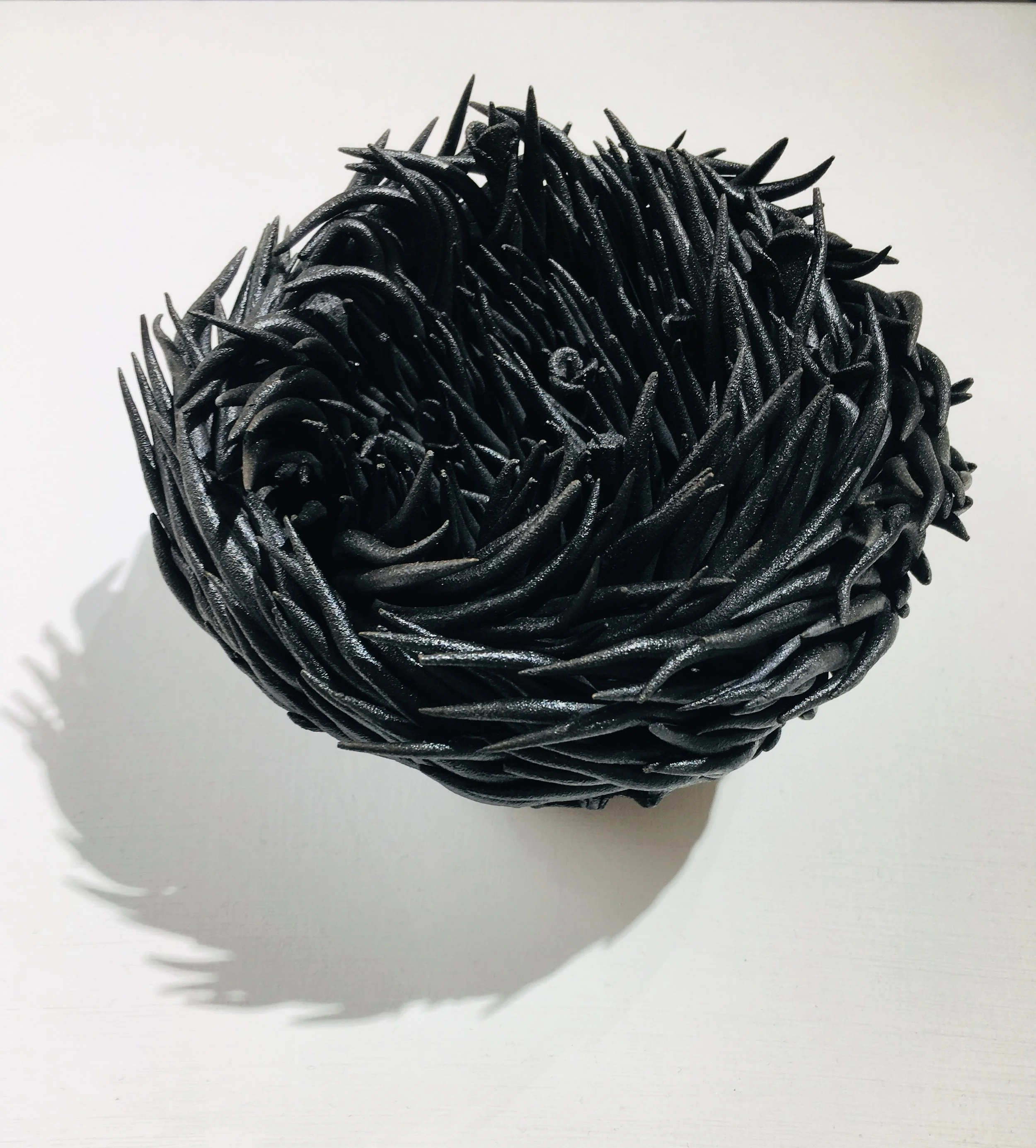 Close-up of a black, spiky, spherical sculpture or object made of intertwined and pointed material, placed on a white surface with shadows.