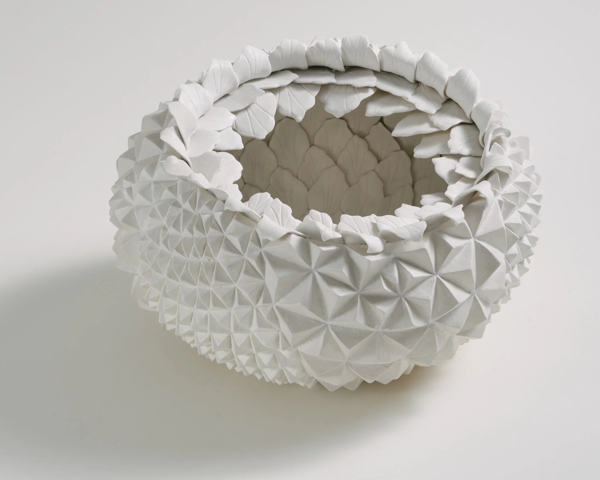 A white ceramic vase with a textured geometric surface and a design of layered, raised leaves around the opening.