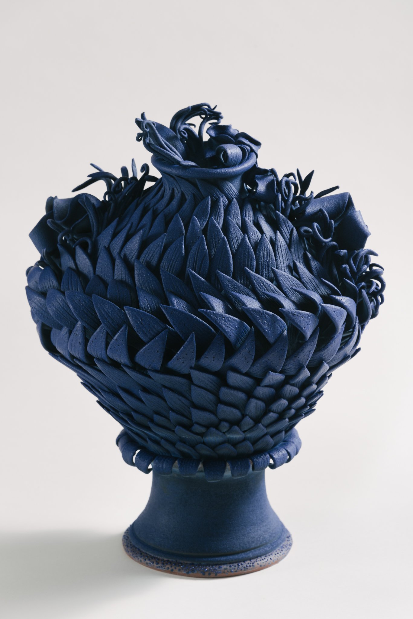 A decorative ceramic vase with a textured, braided design, painted in shades of blue, displayed on a pedestal against a plain white background.