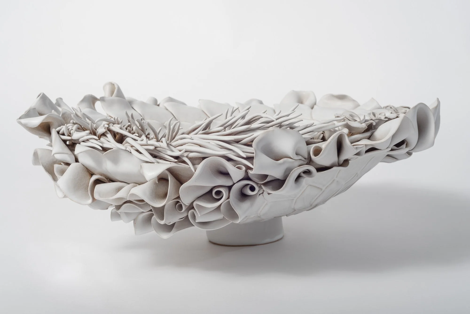 A decorative white ceramic bowl with an intricate design resembling ruffled petals, filled with white seashells, on a plain white surface against a plain white background.