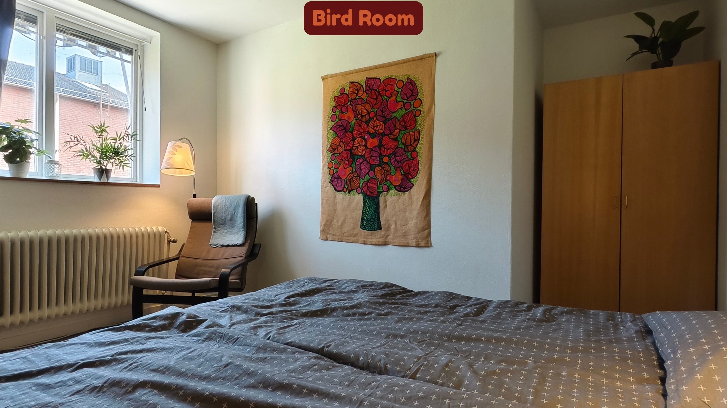 A cozy bedroom with a large window, a potted plant on the sill, a desk lamp, a comfortable chair with a towel draped over it, a colorful fabric wall hanging with a tree design, a wooden wardrobe, and a bed with a patterned blanket.