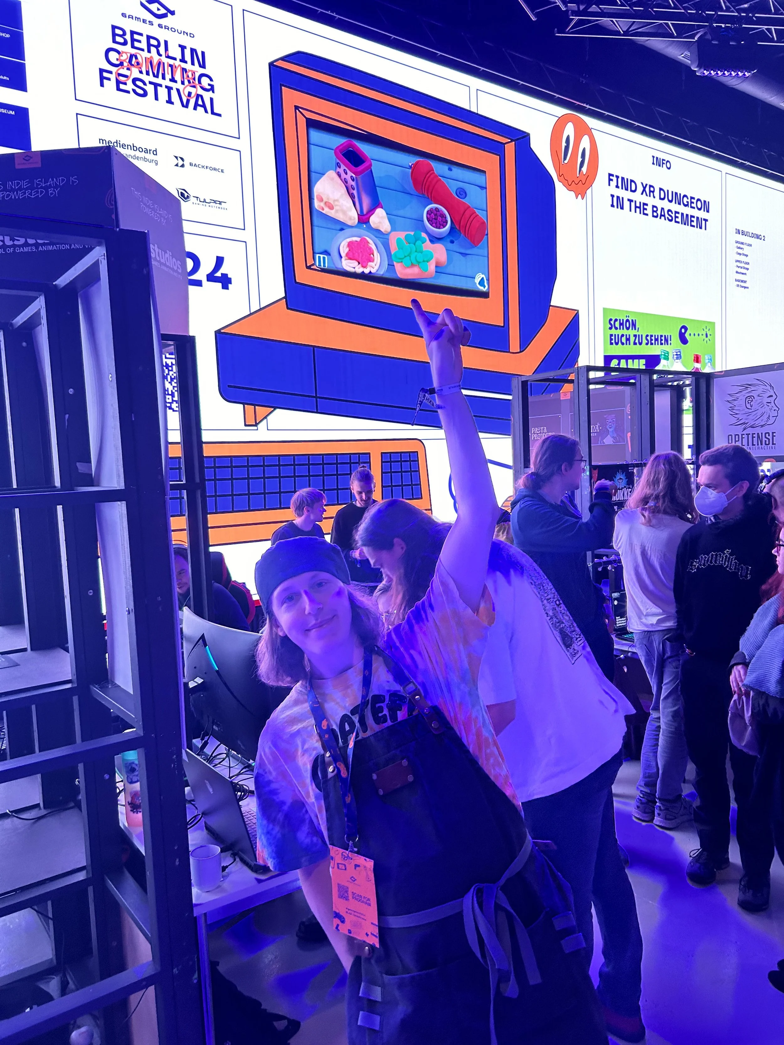 Harry pointing up at his game Pawsta on the big tv at Berlin Games Ground. The room and lighting is very purple.
