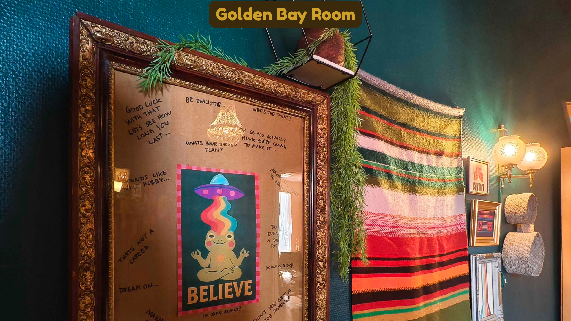 Decorated interior wall labeled 'Golden Bay Room' with framed artwork, handwritten notes, a map, a hanging plaid textile, greenery, and a wall lamp with two glass globes.