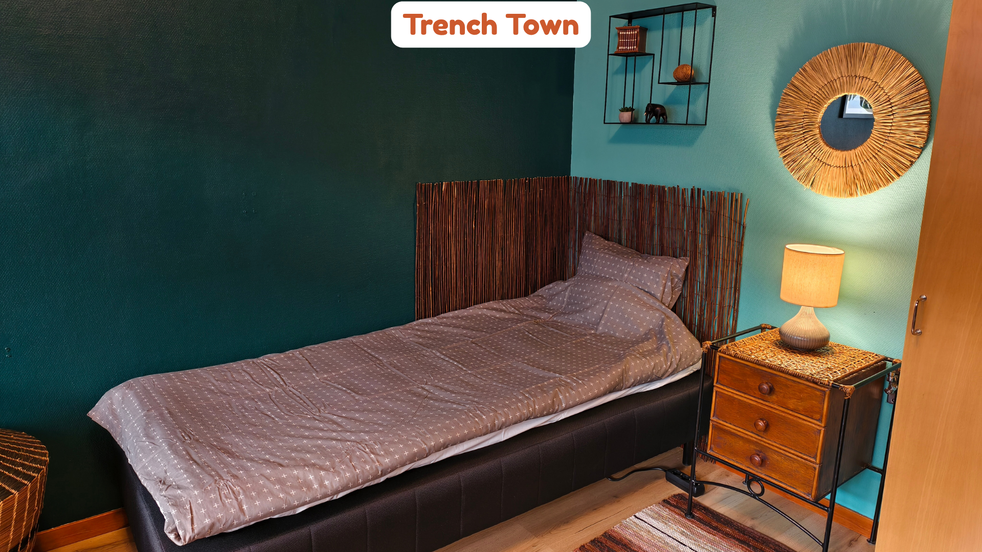 A bedroom with a bed featuring a rattan headboard and light brown bedspread, a wooden nightstand with a lamp, a teal-painted wall with a round straw mirror, a black metal shelf with plants and decor, and a wooden wardrobe door.