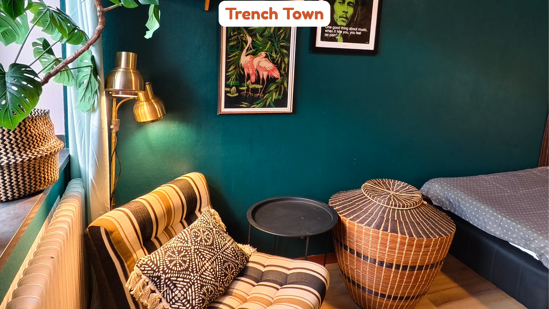 Cozy interior with green walls decorated with framed artwork including a flamingo painting and a portrait, a striped chair with patterned pillows, wicker side tables, a bed with star-printed bedsheet, a tall green plant, and warm lighting.