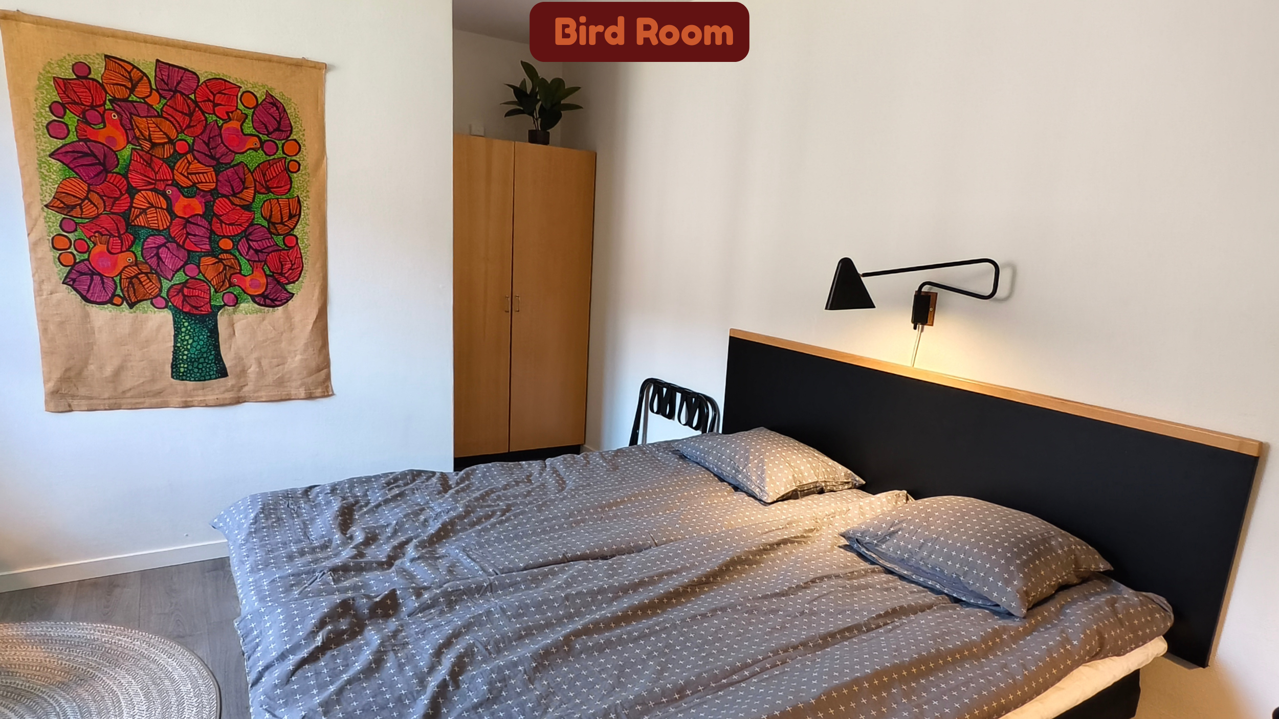 A bedroom with a bed that has gray bedding with white star patterns, a black headboard, and a black wall-mounted light above. To the left of the bed is a wooden wardrobe with a potted plant on top. On the wall above the bed is a red sign that says 'B