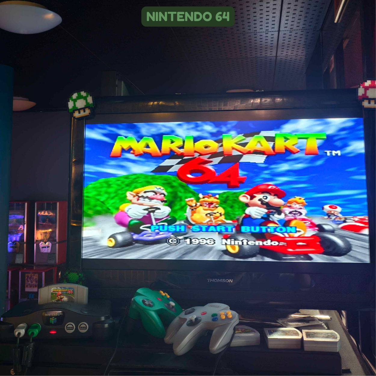 Screenshot of the Nintendo 64 game Mario Kart 64 on a television screen, with controllers and game cartridges in front. The game menu shows characters like Mario, Luigi, Princess Peach, Toad, and Donkey Kong, on a colorful track background.
