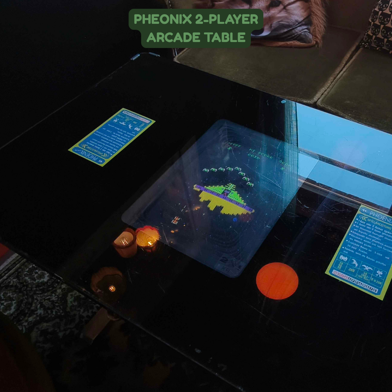 A black arcade table with a glass touchscreen displaying a vintage arcade game. There are two blue and yellow informational stickers on the table. Part of a sofa with a dog print pillow is visible in the background.