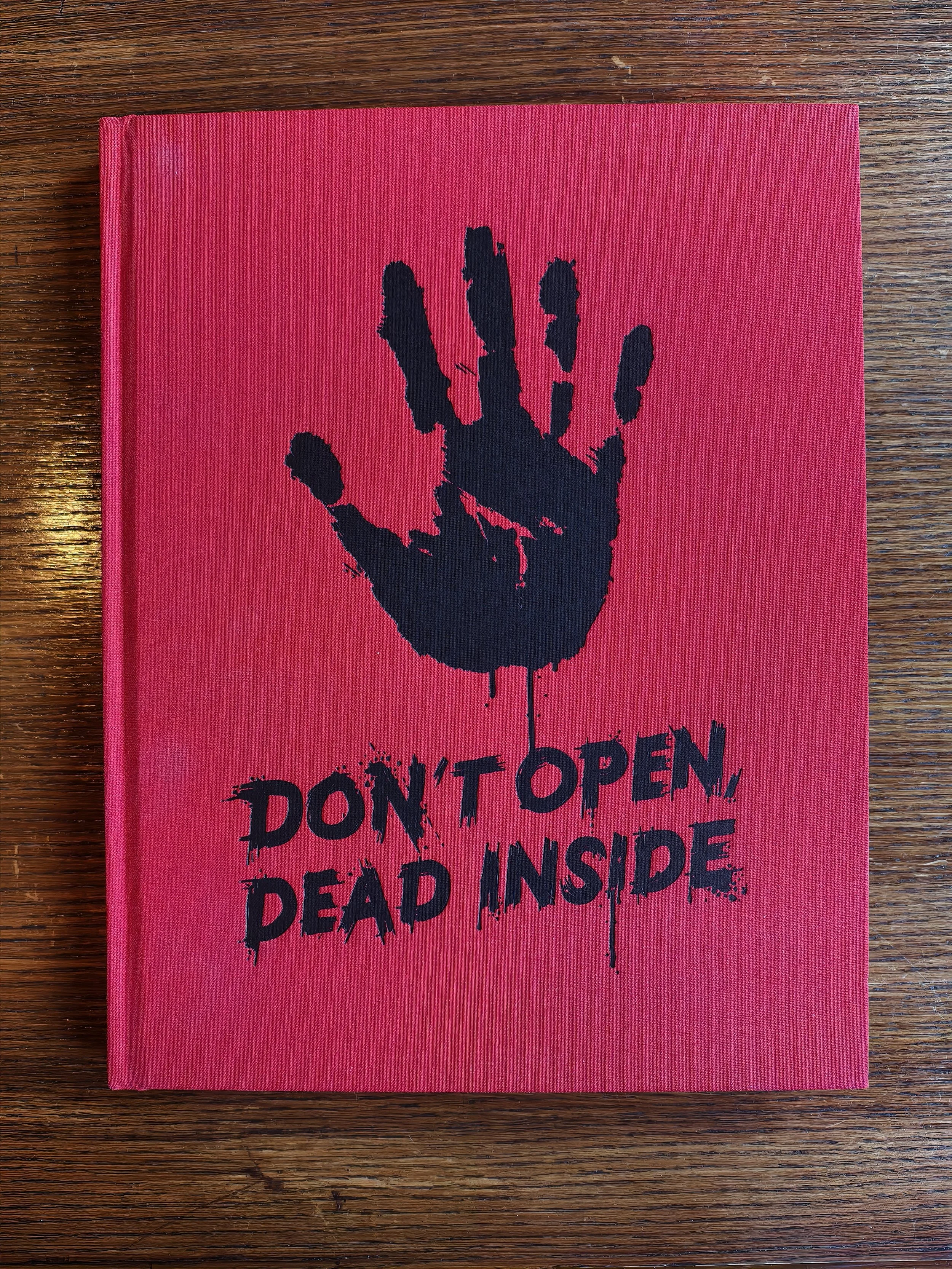 Roleplaying book for The Walking Dead featuring a black handprint on the cover and the words "Don't Open Dead Inside" written below it, placed on a wooden surface.