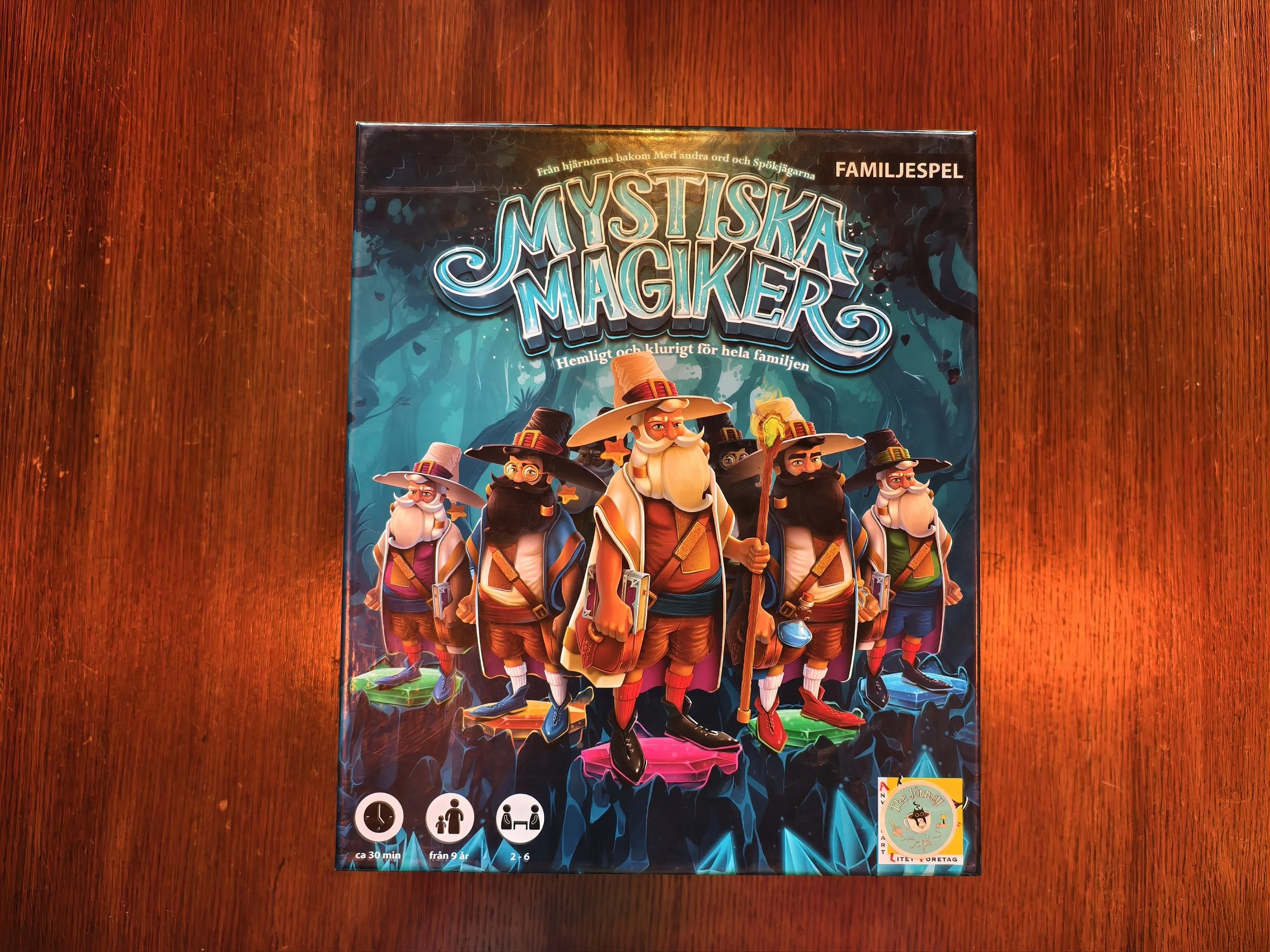 A colorful family board game box titled 'Mystiska Magiker' features animated wizard characters in a fantasy forest setting on a wooden surface.