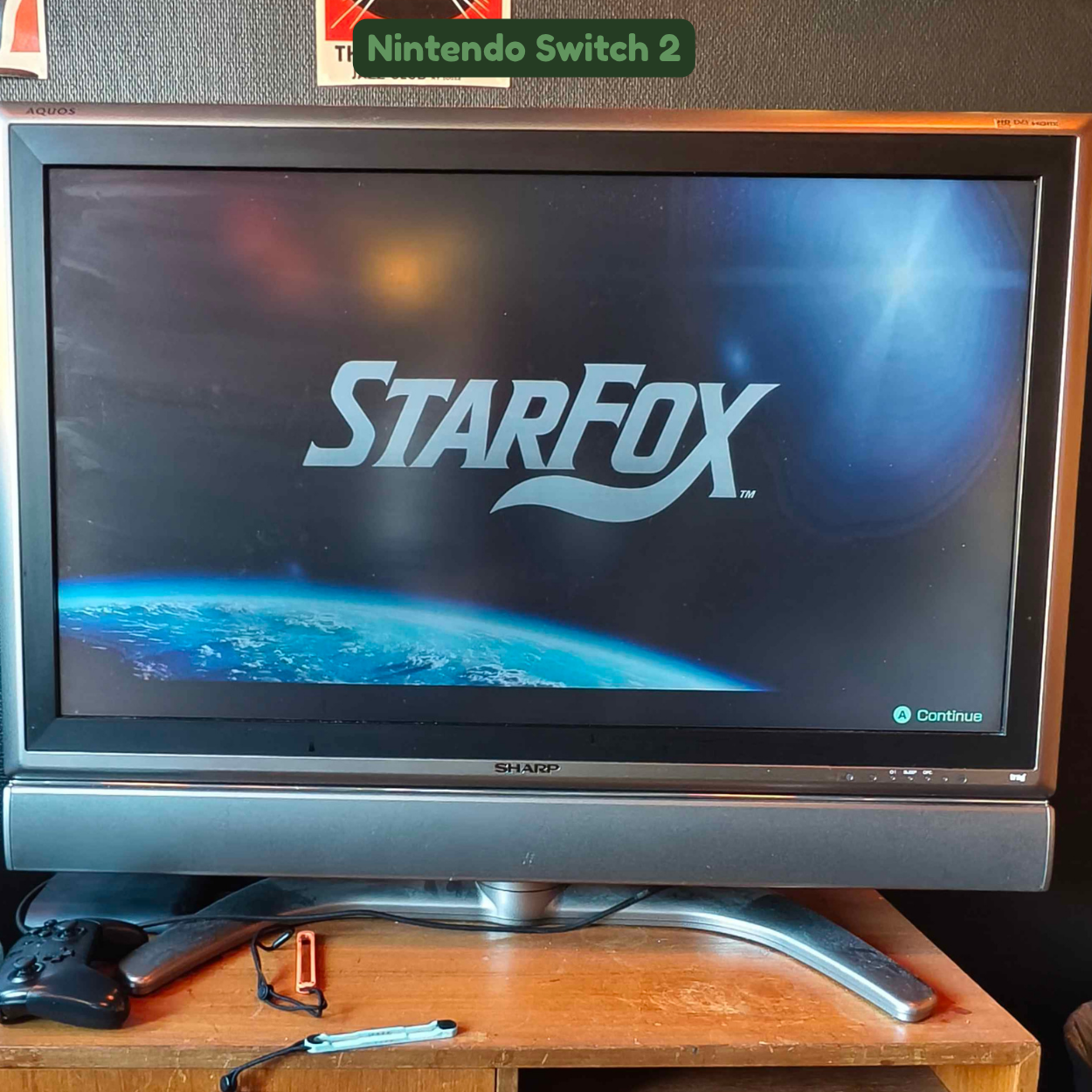 A television screen displaying the title screen for the game 'Star Fox'. The background shows Earth from space with the title in the center and an option to continue in the bottom right corner. The TV is on a wooden surface with a game controller, a 
