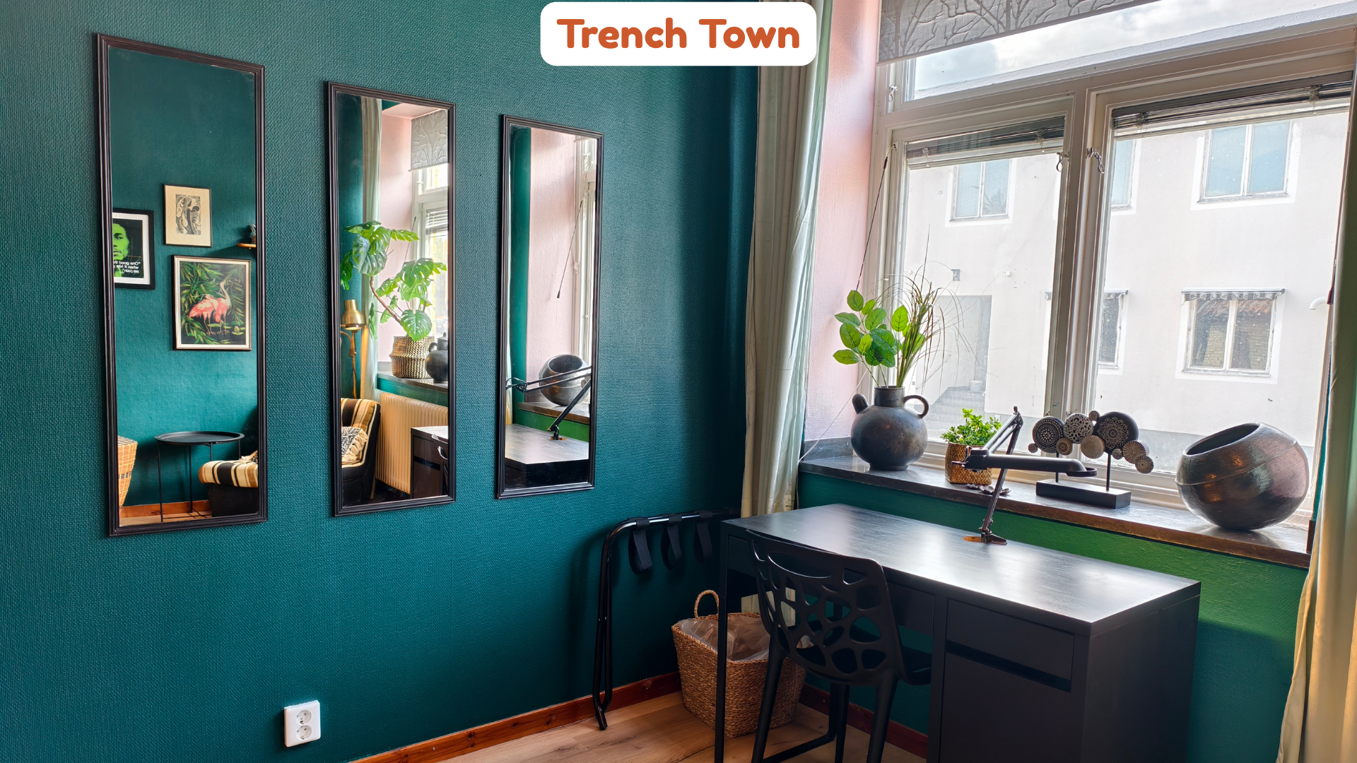 A cozy corner in a room with teal textured walls, decorated with three vertical mirrors and framed artwork. A desk beneath the large windows holds decorative vases with plants, and a chair is tucked underneath. The windows have light-colored curtains