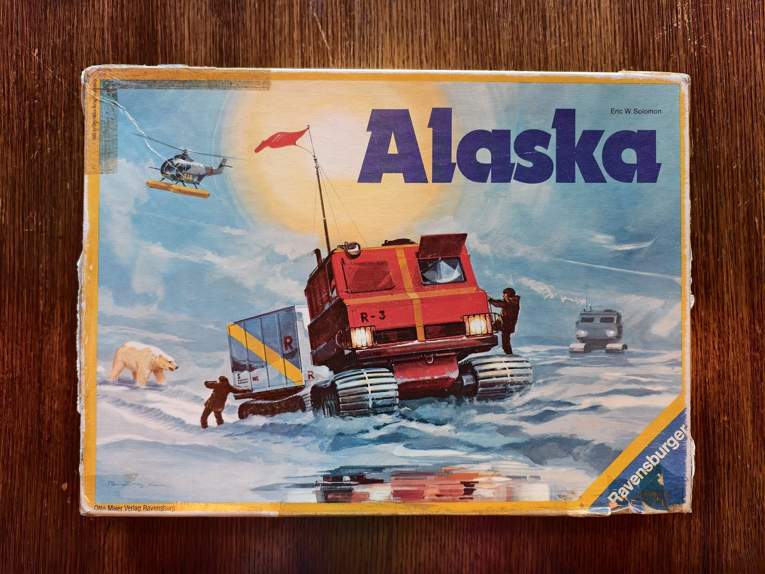 Vintage board game box titled 'Alaska' featuring a red amphibious vehicle, a man helping it across snow, a helicopter, a dog, and a snowy landscape.