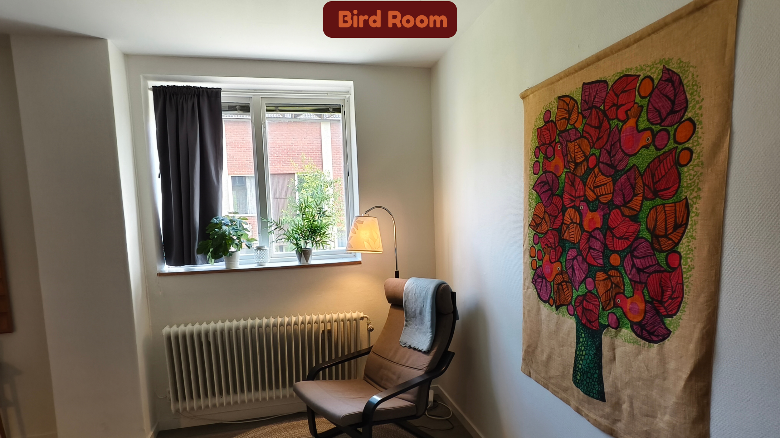 A corner of a room with a window, potted plants, a chair, a lamp, a radiator, and a colorful tapestry of a tree with birds on the wall.