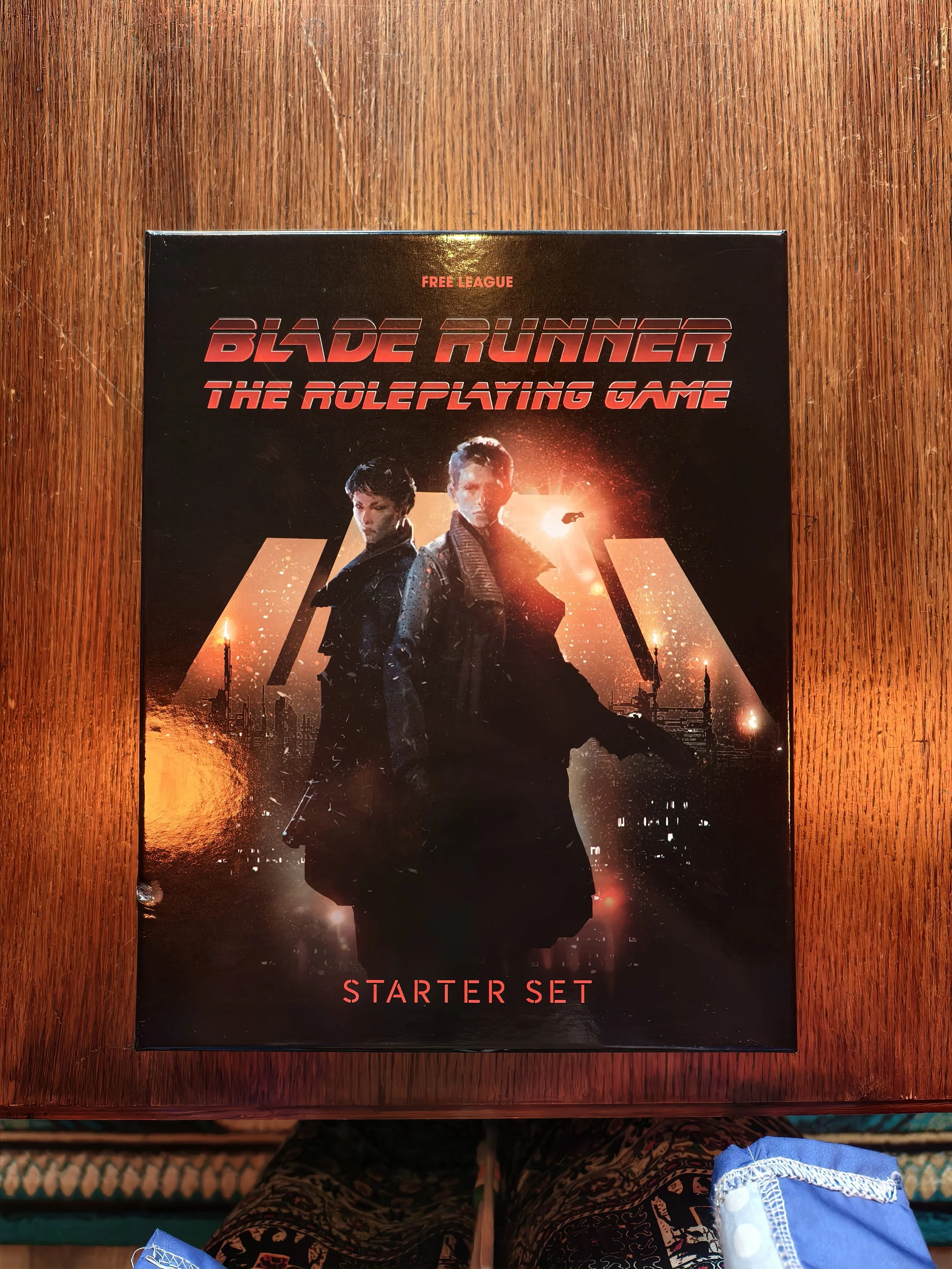 A tabletop board game box titled "Blade Runner: The Roleplaying Game" with a black background, featuring characters from Blade Runner in the foreground, and the words "STARTER SET" in red at the bottom. The table surface is wooden, and part of a pers