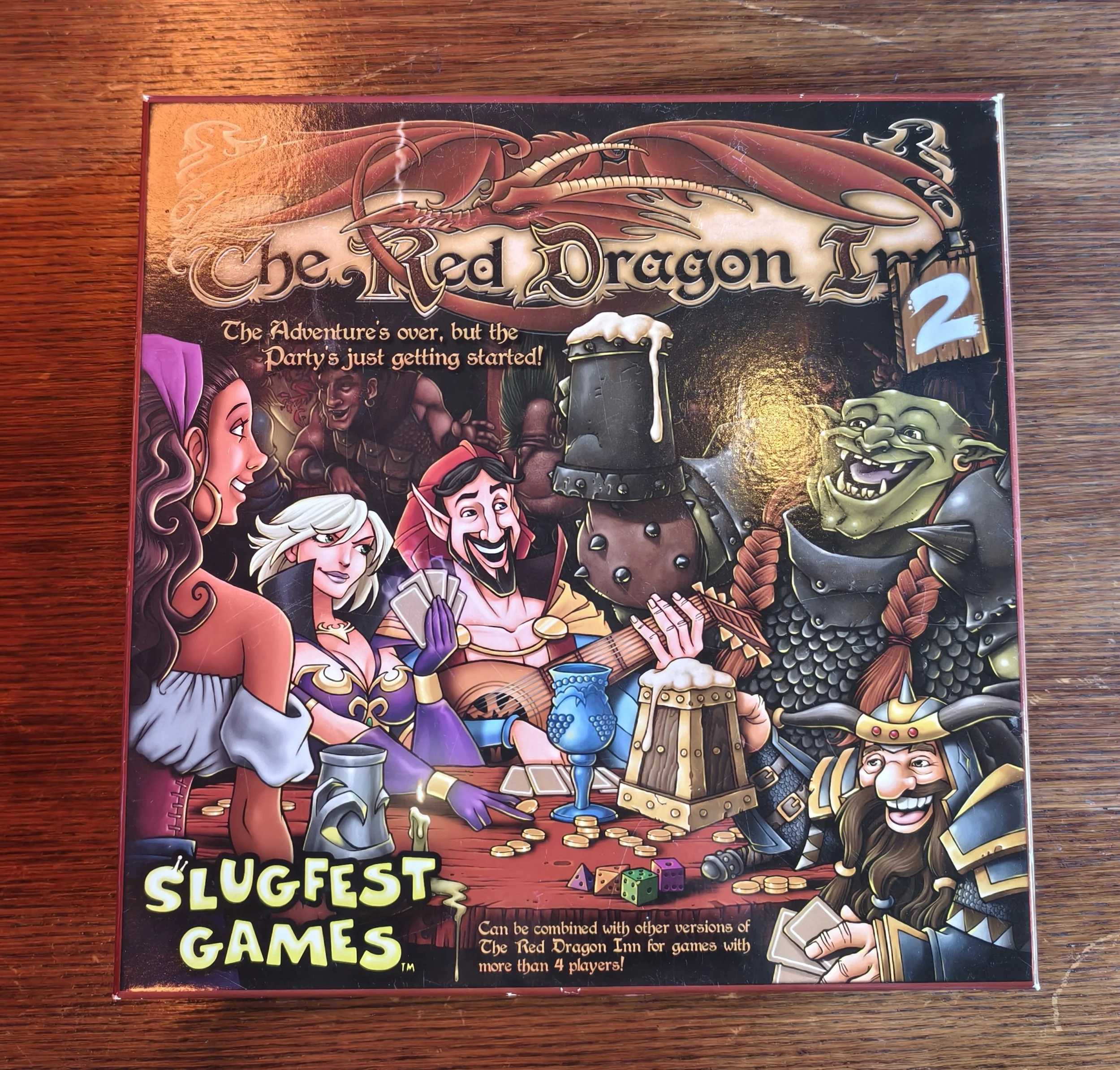 Box cover for a game titled 'The Red Dragon Inn' featuring a cartoon dragon, a pirate, a witch, and a green goblin around a table with dice and coins, on a wooden surface.