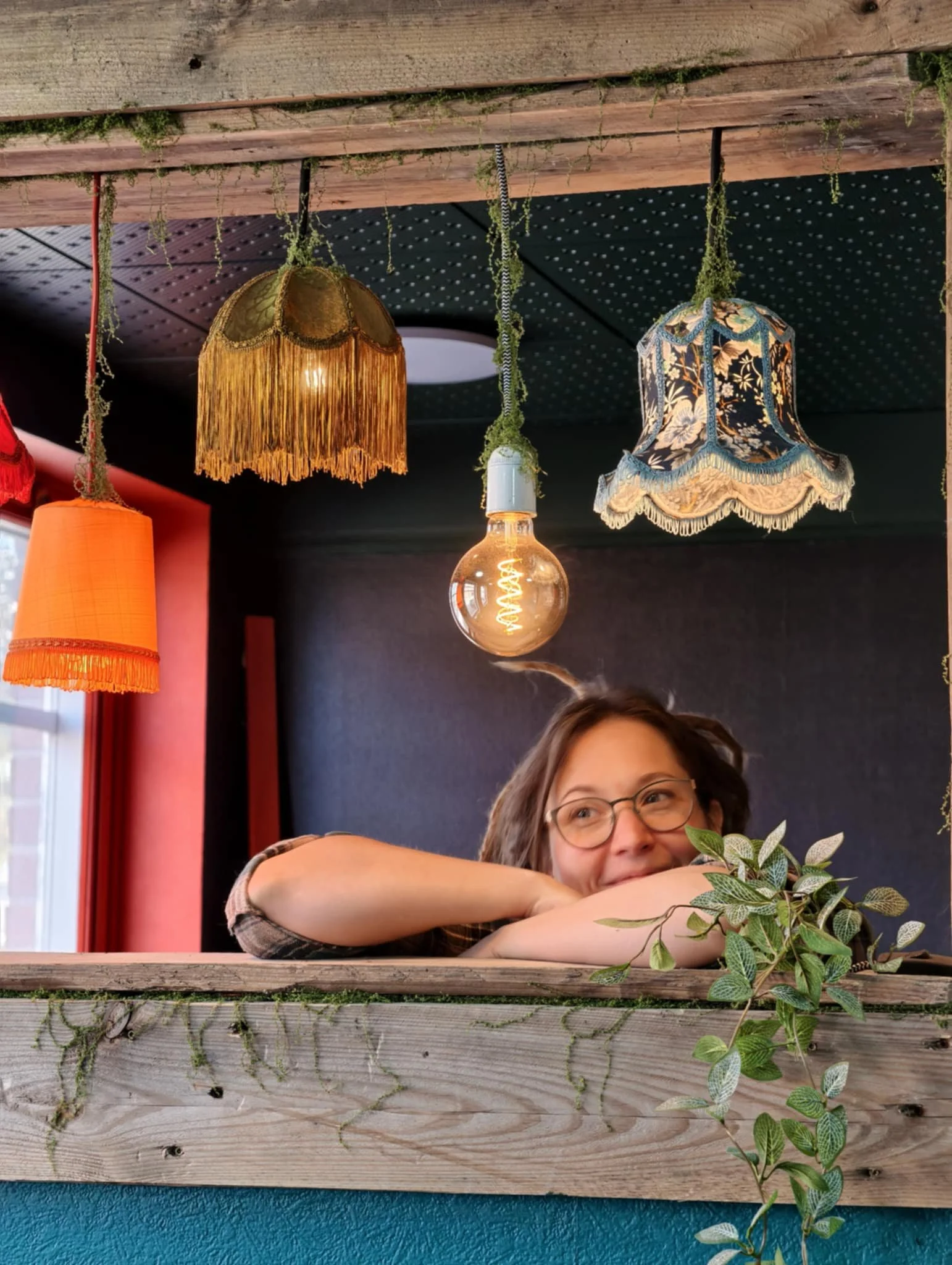 Em at the Jörney Café, gazing through a window. The ceiling has hanging lamps with decorative shades, while the window is made up of repurposed wood, with mossy details added