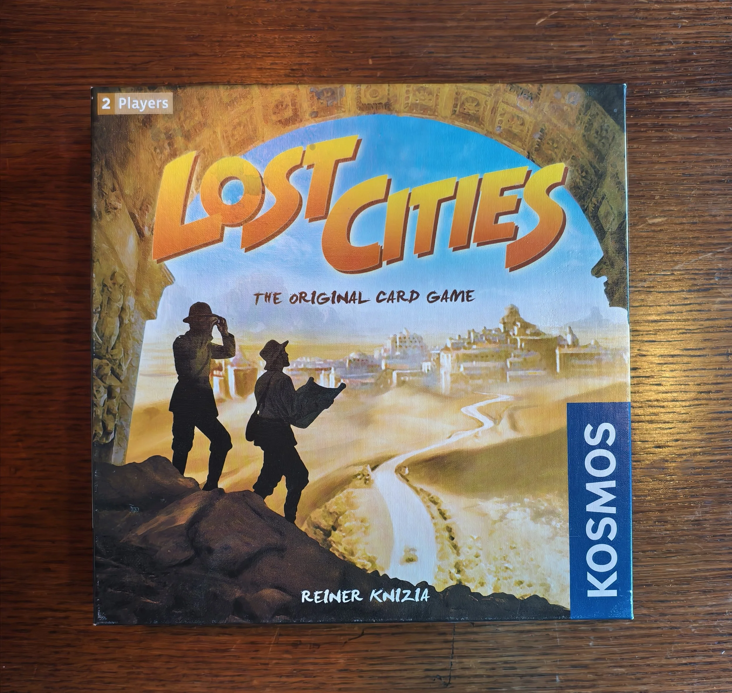 Board game titled 'Lost Cities' with artwork depicting explorers walking on a rocky terrain towards a cityscape at sunset. The game is designed for 2 players, created by Reiner Knizia.