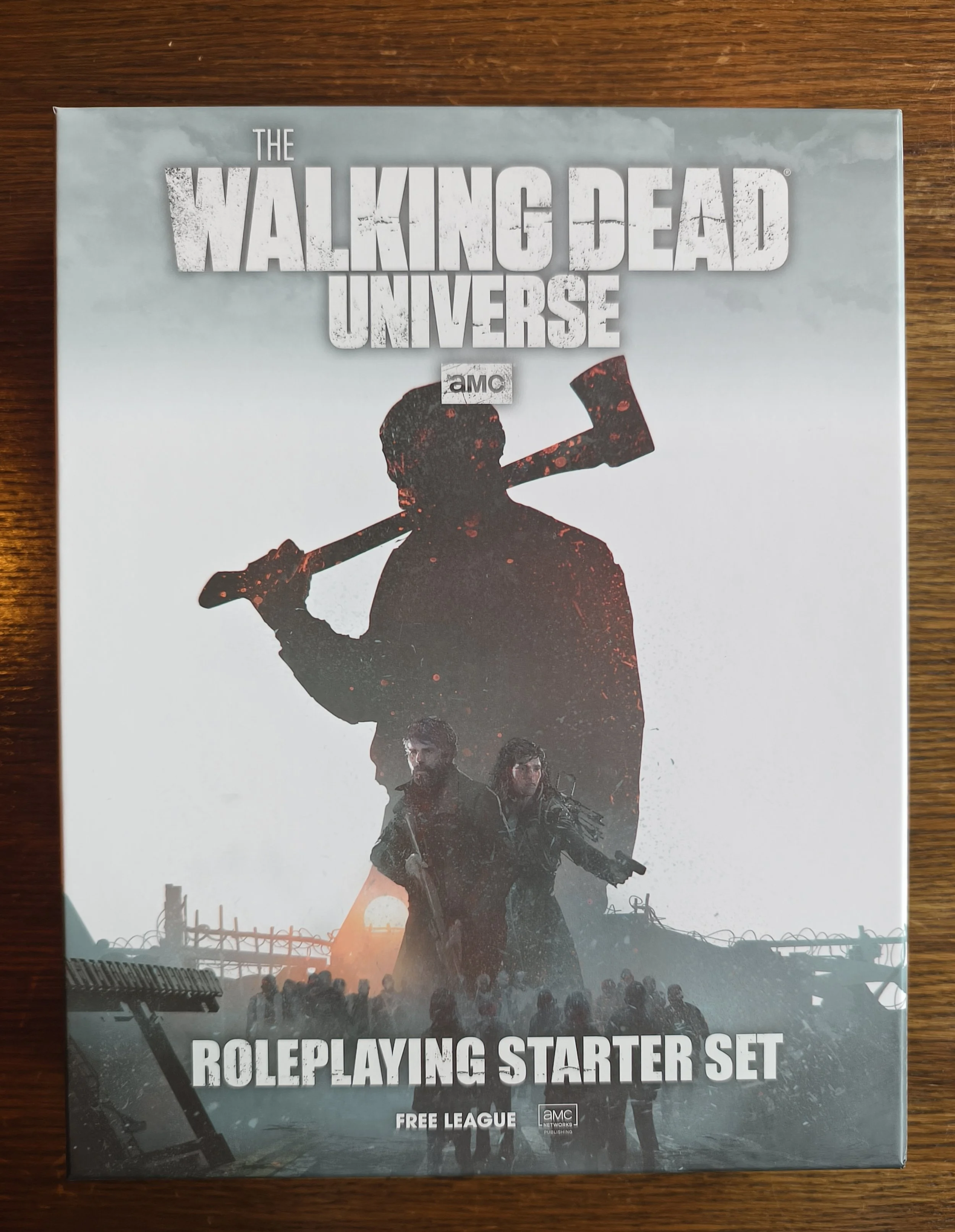 Box art for 'The Walking Dead Universe Roleplaying Starter Set,' featuring silhouettes of armed characters, a barbed wire fence, and a crowd in the background with a sunset sky.