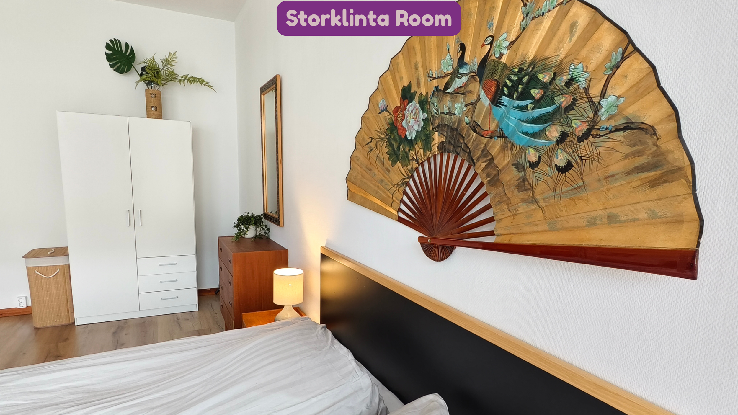 A bedroom with a large decorative fan featuring floral and bird artwork mounted on the wall above a bed with white bedding. There is a bedside table with a small lamp, a mirror on the wall, and a white wardrobe to the side. A small plant is also visi