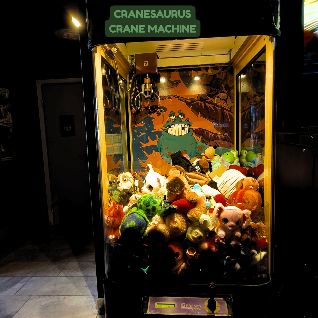 Claw crane arcade game filled with plush toys and stuffed animals, with a cartoon frog illustration in the background.
