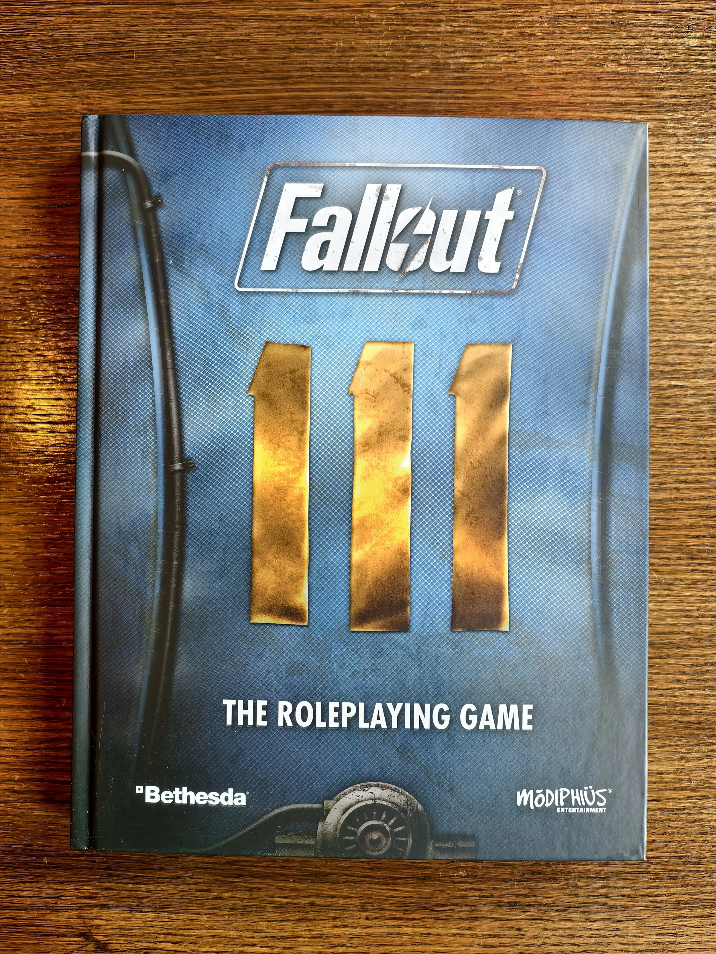 Cover of a role-playing game book titled 'Fallout: The Roleplaying Game' with a blue background, metallic gold numbered '111', and logos of Bethesda and Modiphius Entertainment at the bottom on a wooden surface.
