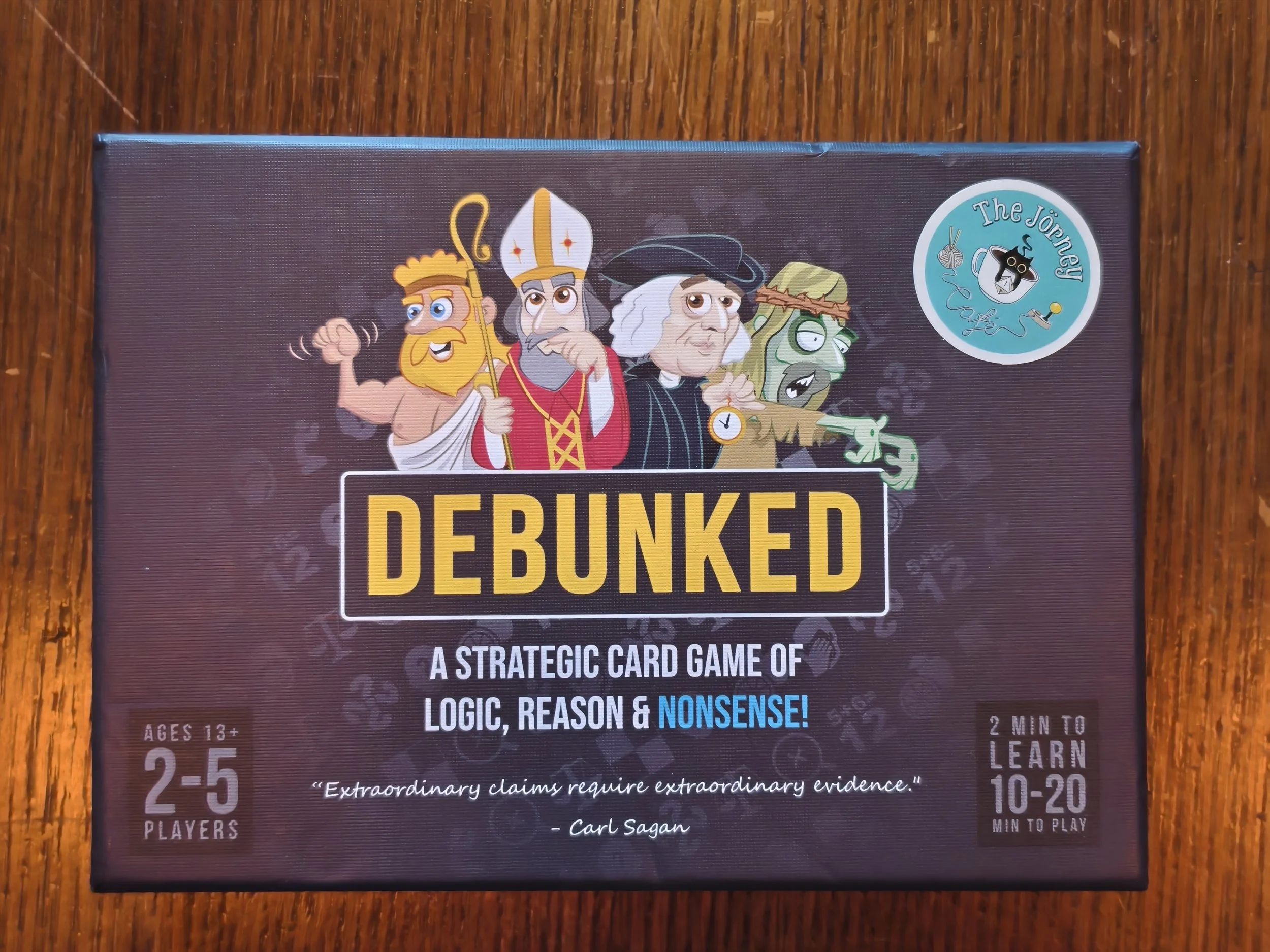 Box of the card game 'Debunked' featuring cartoon characters dressed as a king, wizard, pirate, and witch on the cover. The game is described as a strategic card game of logic, reason, and nonsense for ages 13 and up.