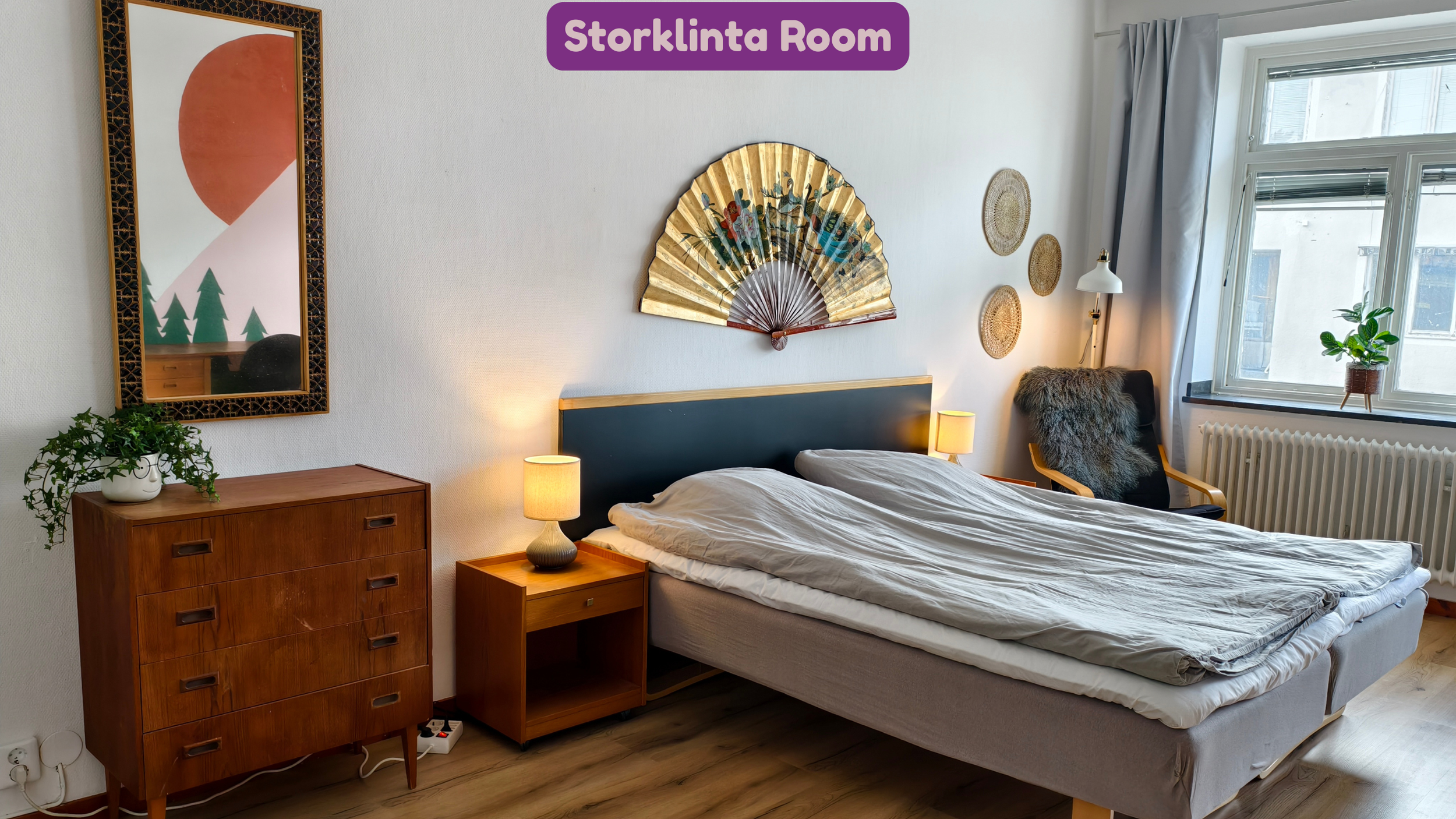 A cozy bedroom with a wooden dresser, a bed with white bedding, and a nightstand with a lamp. Decor includes a fan-shaped wall art, woven baskets on the wall, a large mirror, a potted plant, and a window with a curtain, all labeled 'Storklinta Room'.