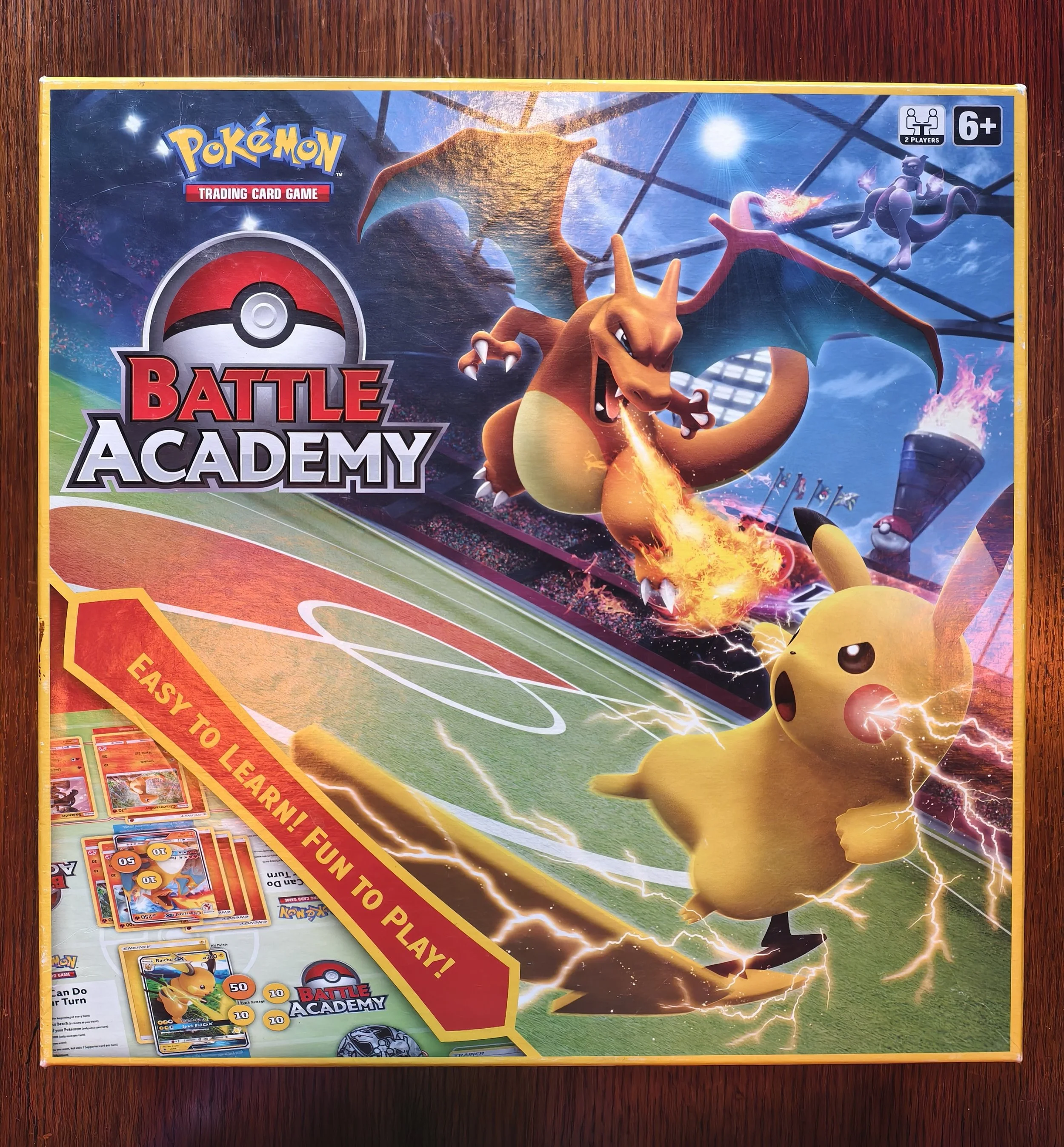 Pokémon Battle Academy trading card game box with Charizard, Mewtwo, Pikachu, and various Pokémon cards on a wooden surface.