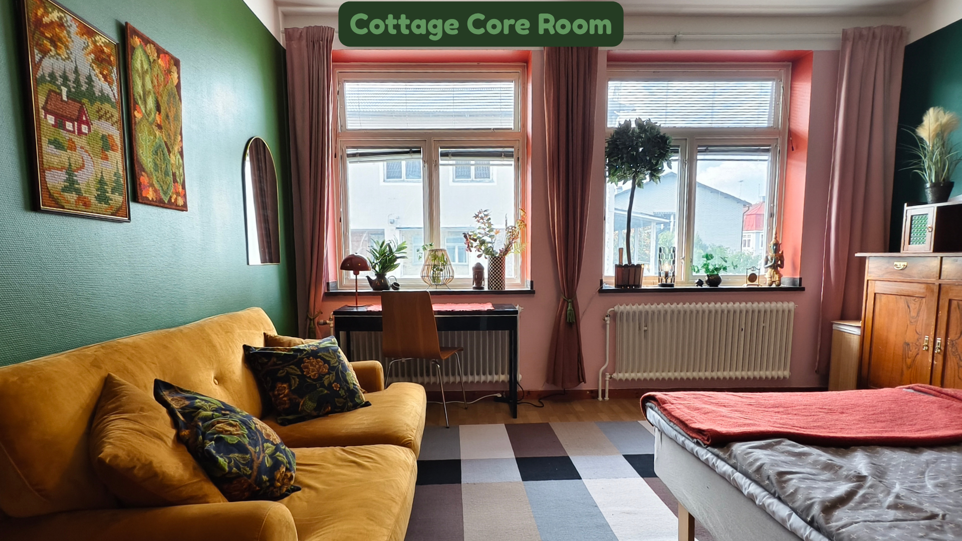A cozy room with green and pink walls, a mustard yellow sofa with patterned pillows, two large windows with pink curtains, a small desk with plants and decorative items, a radiator, a wooden cabinet with decorative objects, and a bed with a red blank