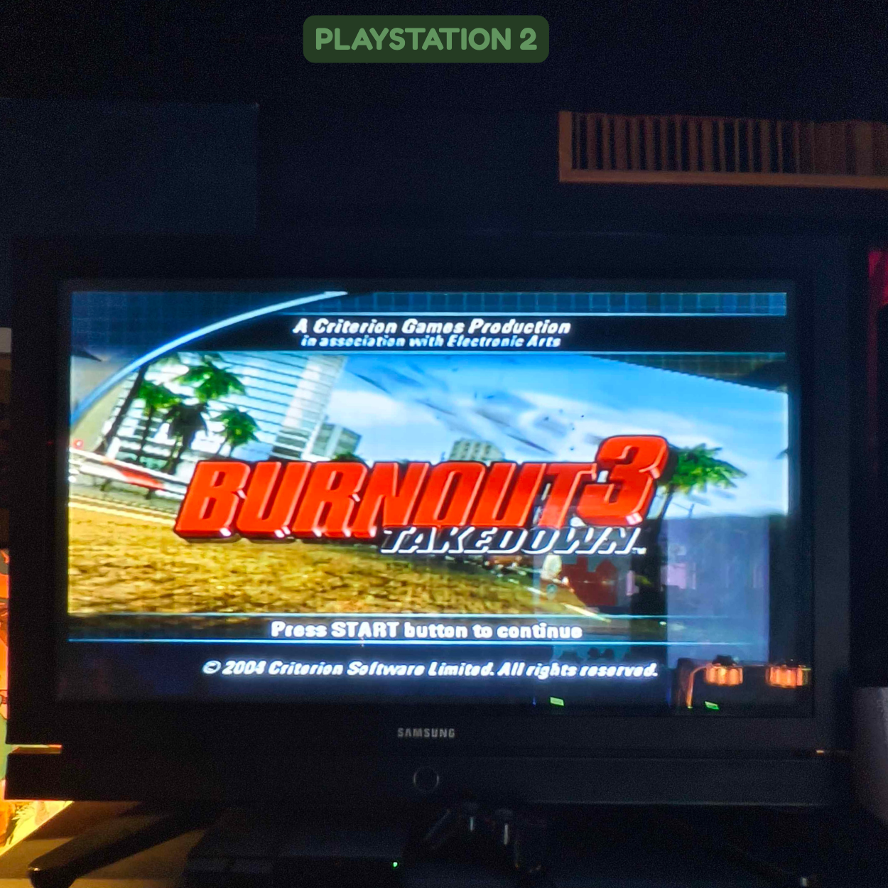 Title screen of Burnout 3 Takedown video game on a Samsung monitor, with cityscape background and palm trees, displaying the message to press start to continue.
