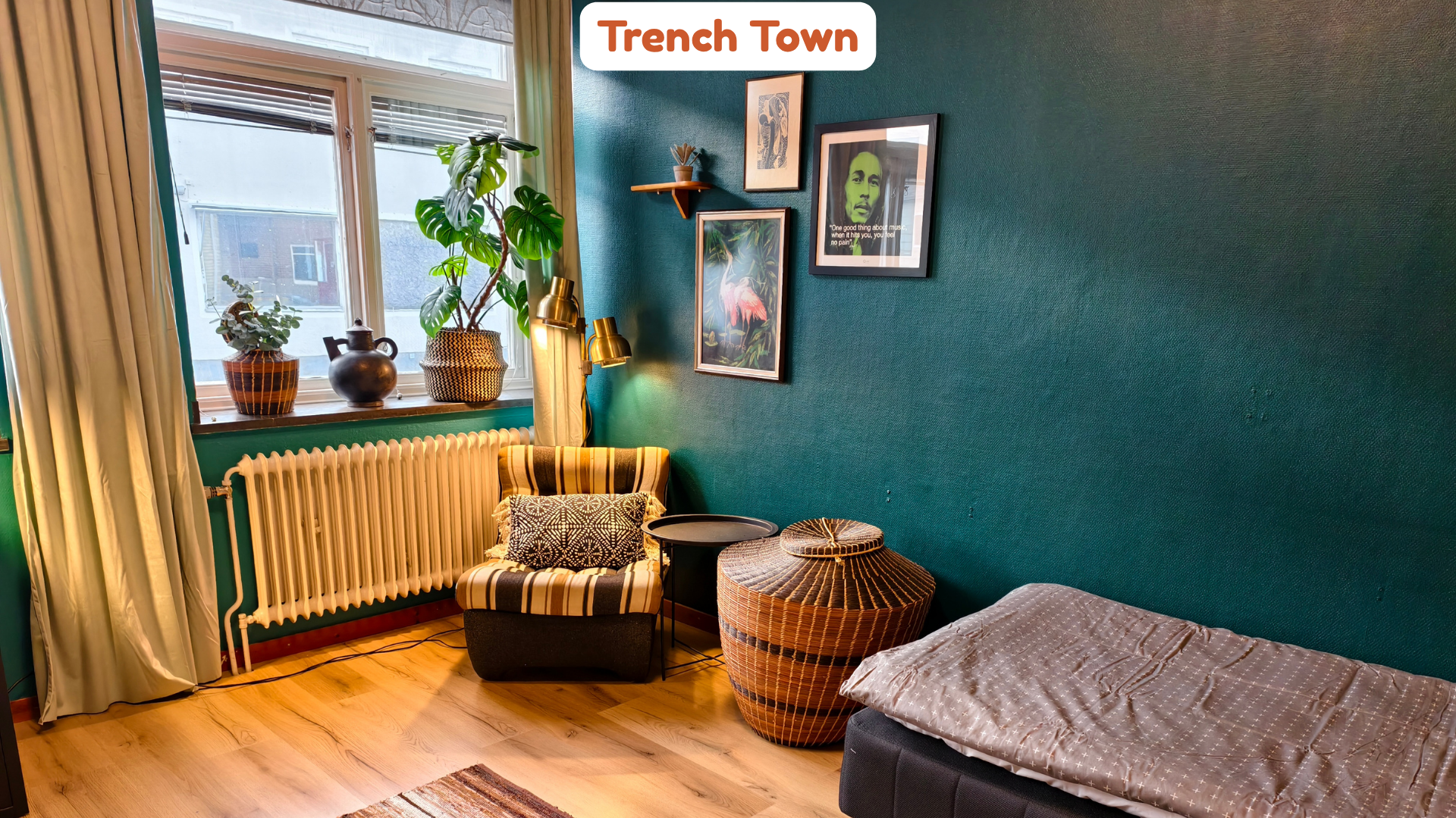 A cozy indoor space featuring a window with green curtains, plants on the windowsill, framed artwork on a teal wall, a striped armchair with a patterned pillow, a small black side table, a woven basket, a bed with a light-colored blanket, and a white