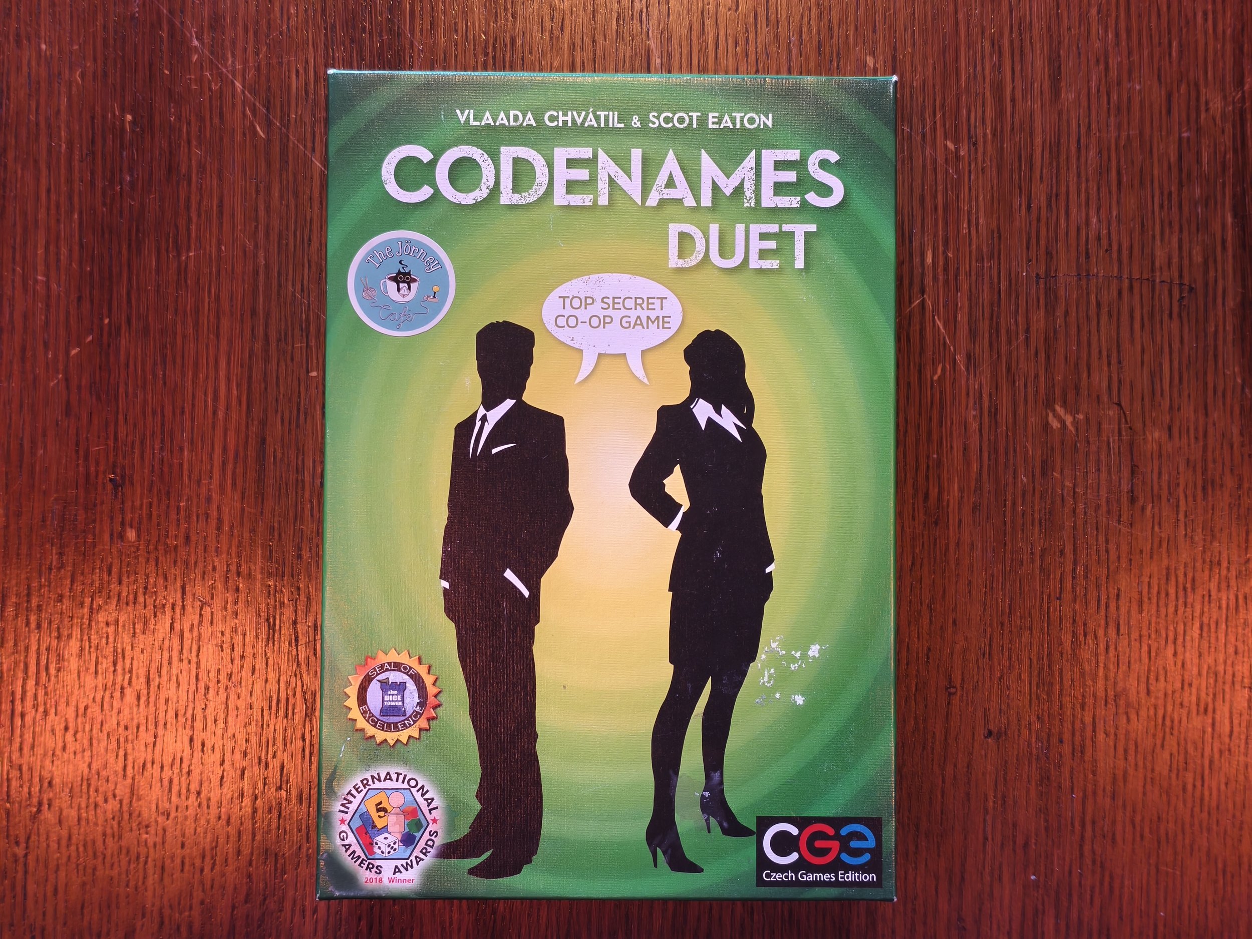 Cover of the game 'Codenames Duet' with a green background, featuring black silhouettes of a man and a woman in business attire facing each other, talking with a speech bubble. The cover includes logos indicating awards and editions, such as 'Czech G