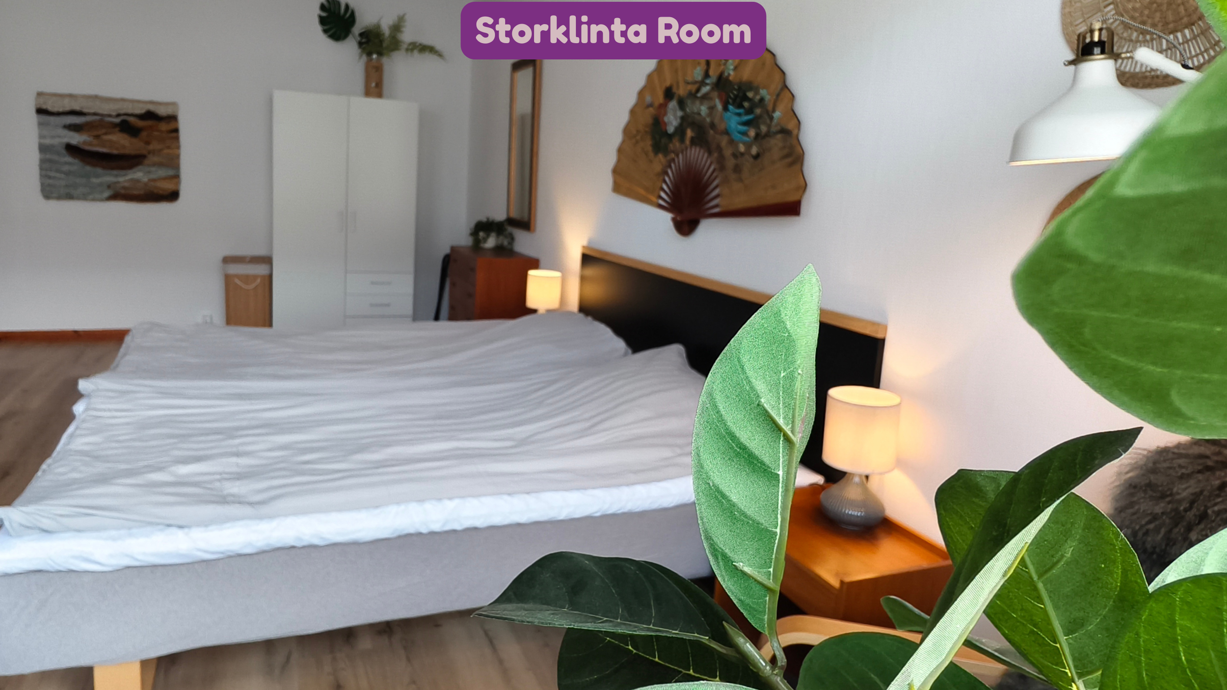 A bedroom with a bed covered in white sheets, a white wardrobe, a wooden nightstand with a small lamp, a decorative wall fan, and plants, creating a cozy and natural ambiance.
