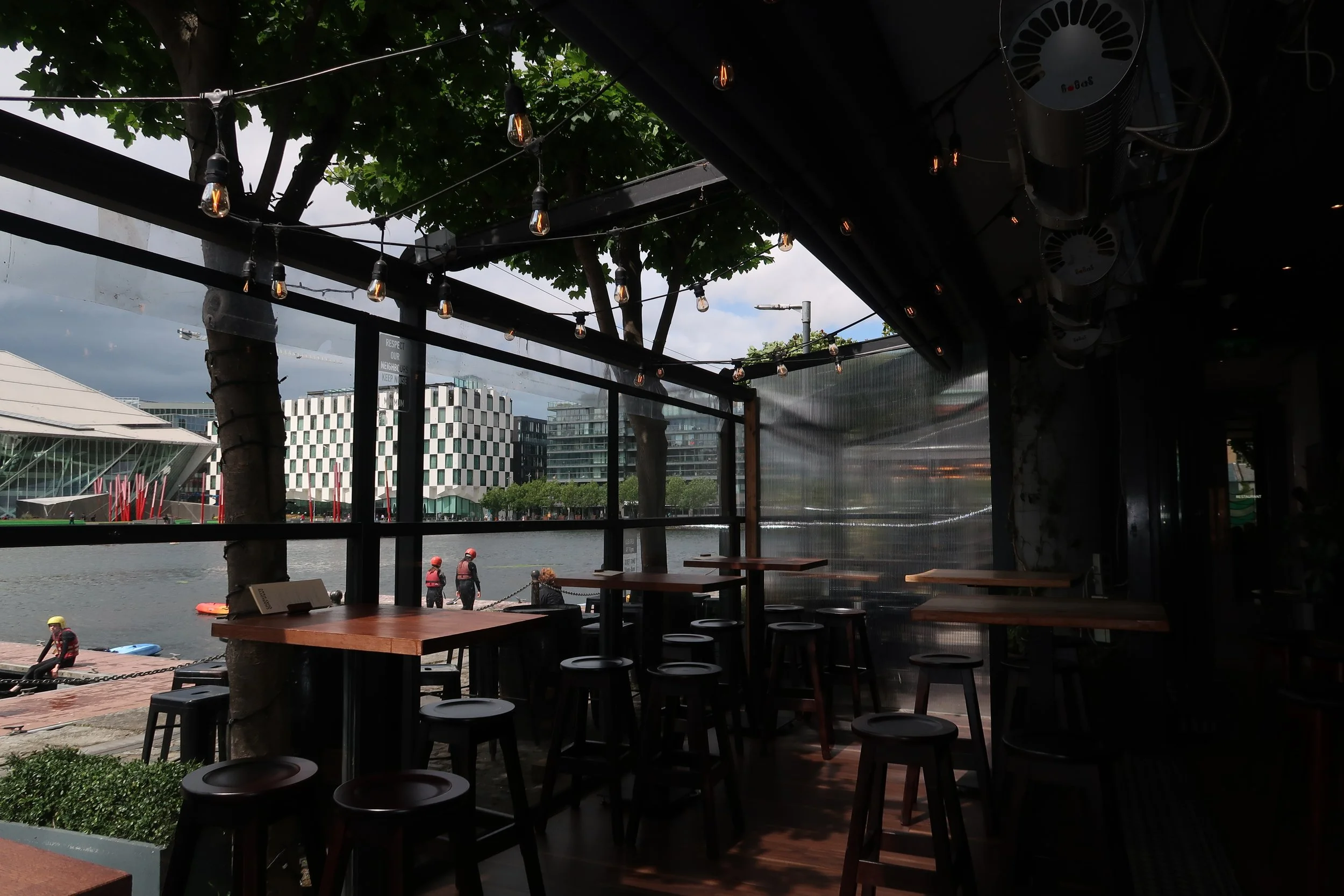 Interior of a restaurant or cafe with wooden tables and stools, overlooking a river with people in life vests near the water, trees and modern buildings in the background, and string lights hanging from the ceiling.